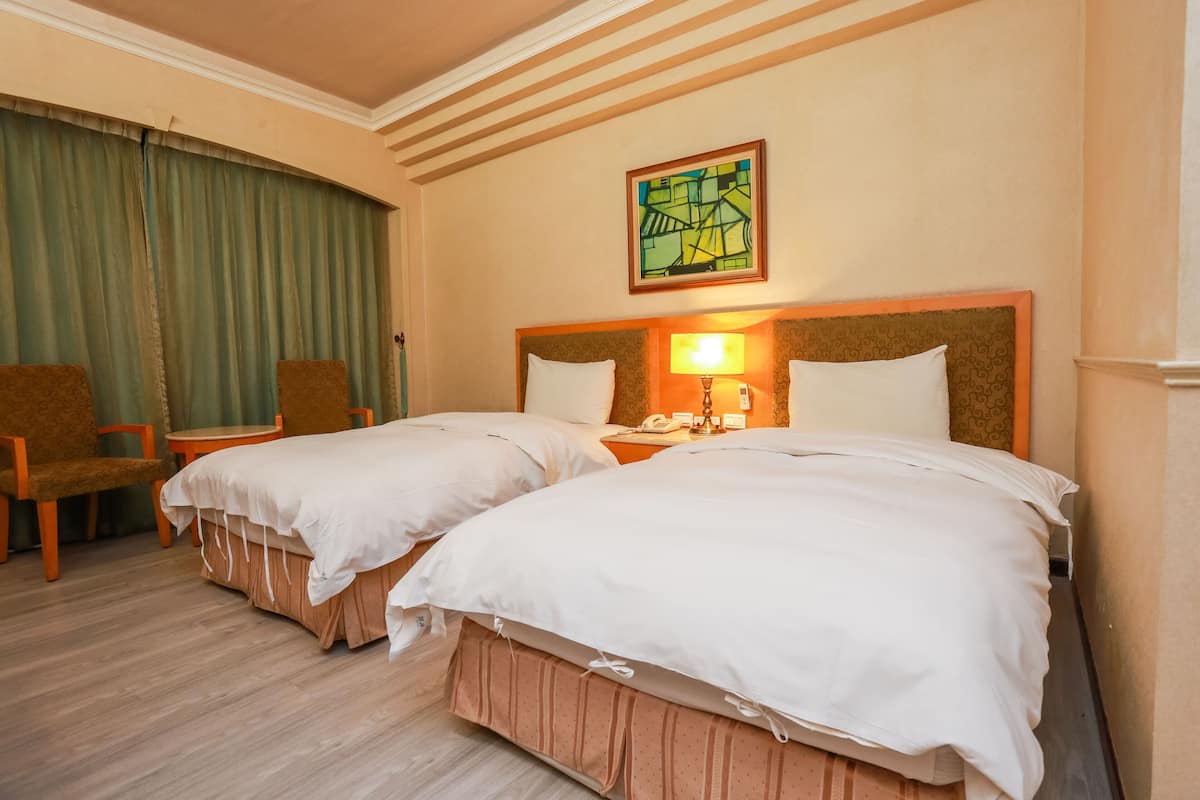 A hotel room with two beds, a desk, chairs, a lamp, and a painting on the wall.