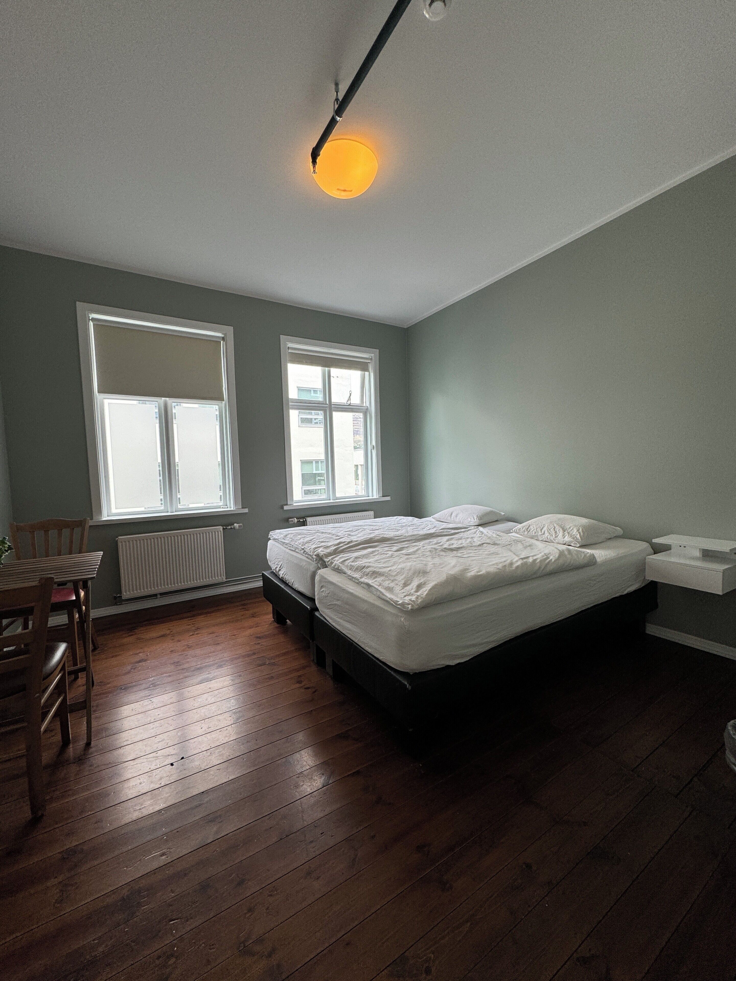 Un dormitorio con cama, una silla, una mesa y dos ventanas.