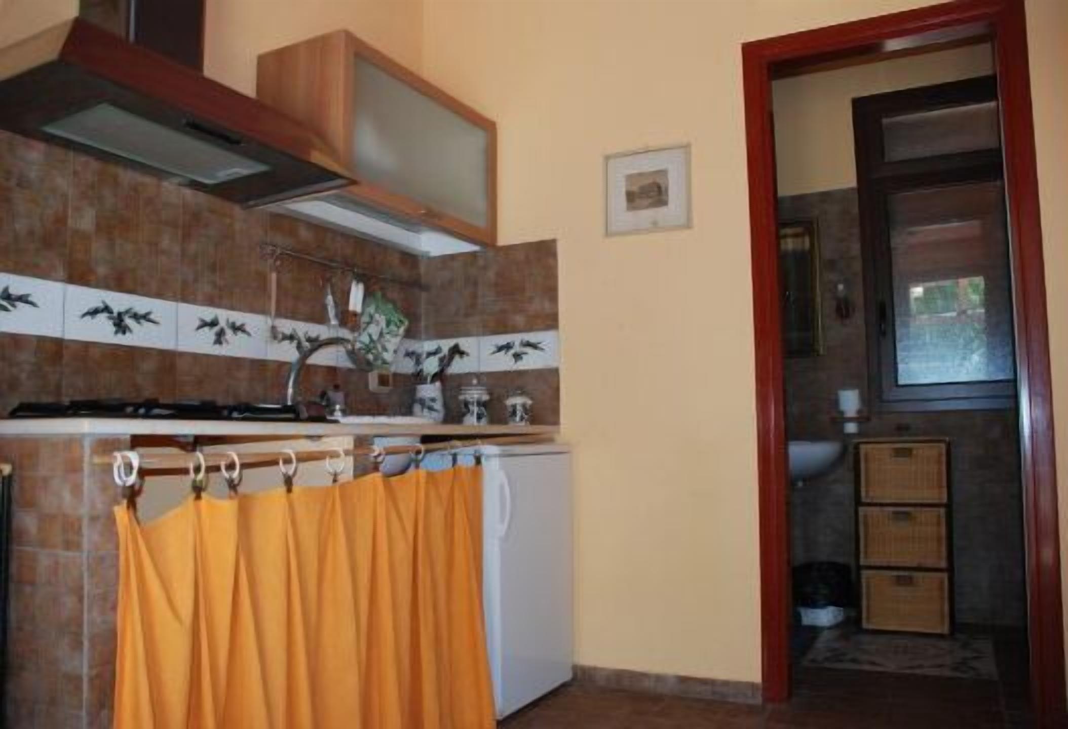 Cocina compacta con estufa, lavamanos y un refrigerador pequeño. Hay un gabinete de madera sobre la estufa y un protector de salpicadura de azulejos.
