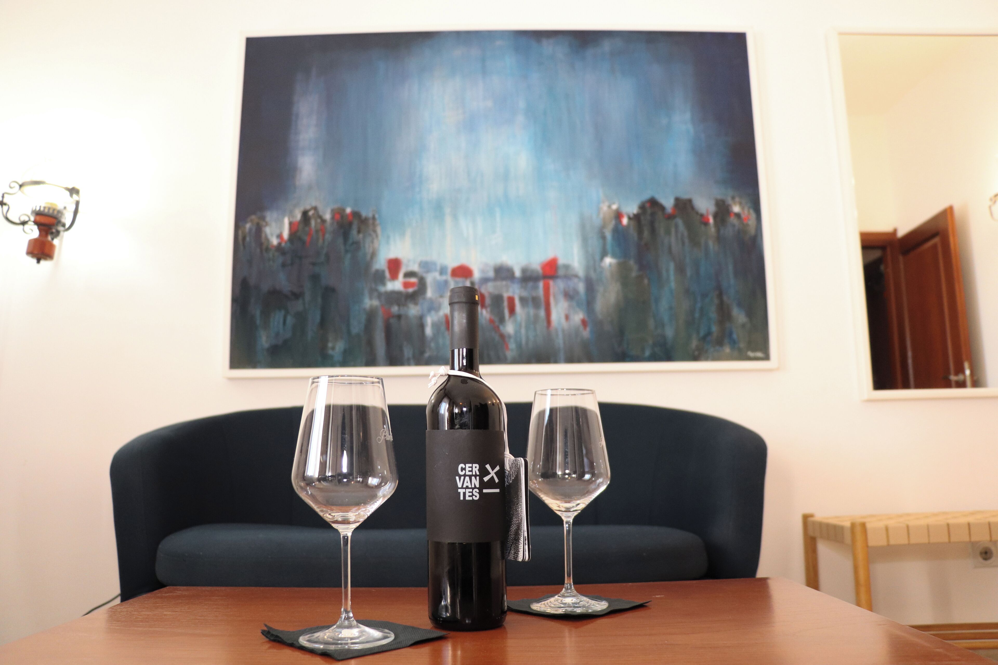 Una mesa con una botella de vino y dos copas, una silla azul oscuro y un cuadro en la pared.