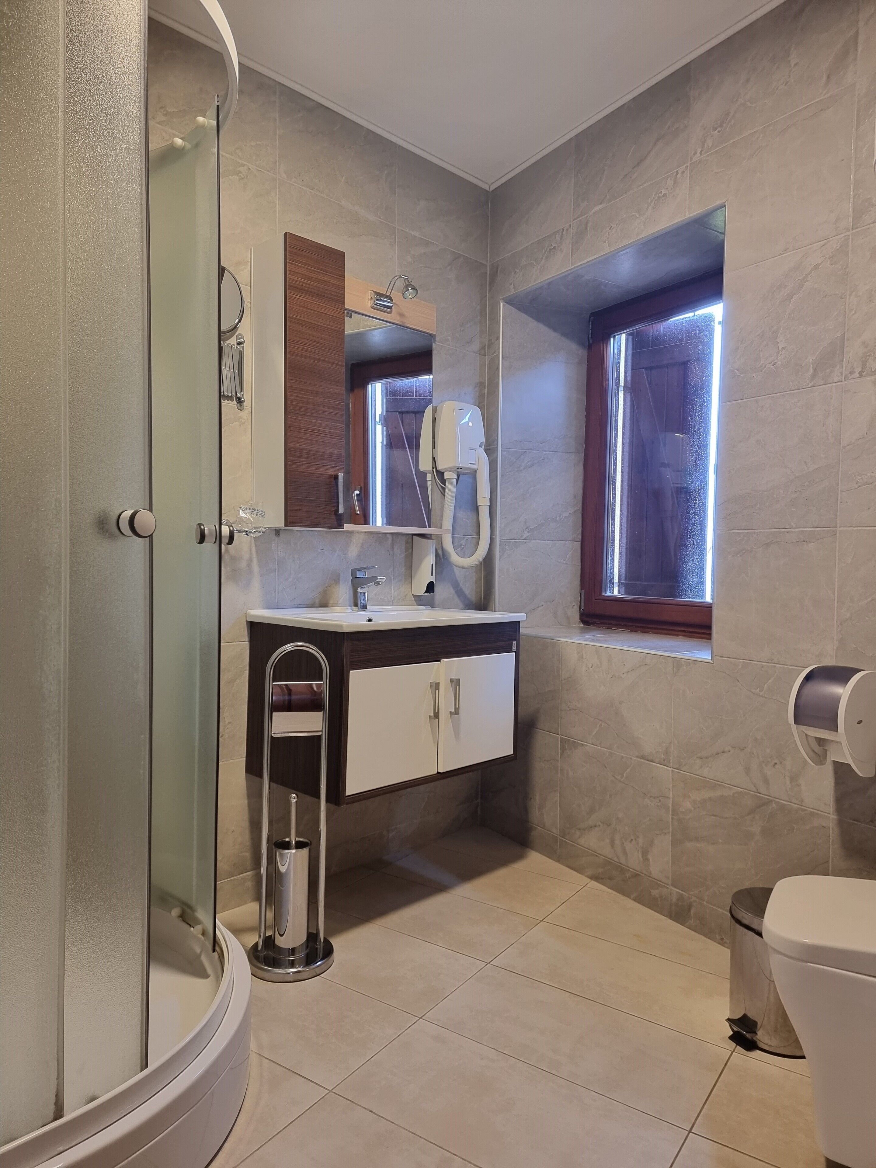 Un baño moderno con cabina de ducha de vidrio, un lavabo blanco con mueble de madera, un inodoro y una ventana que proporciona luz natural.