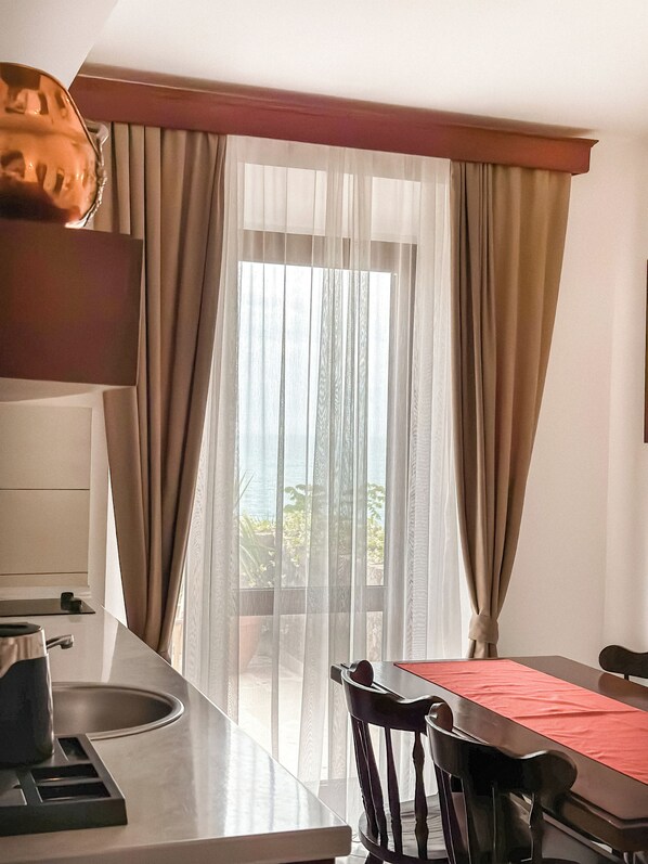 A hotel room with a dining area, a large window with sheer curtains, and a view of greenery outside.