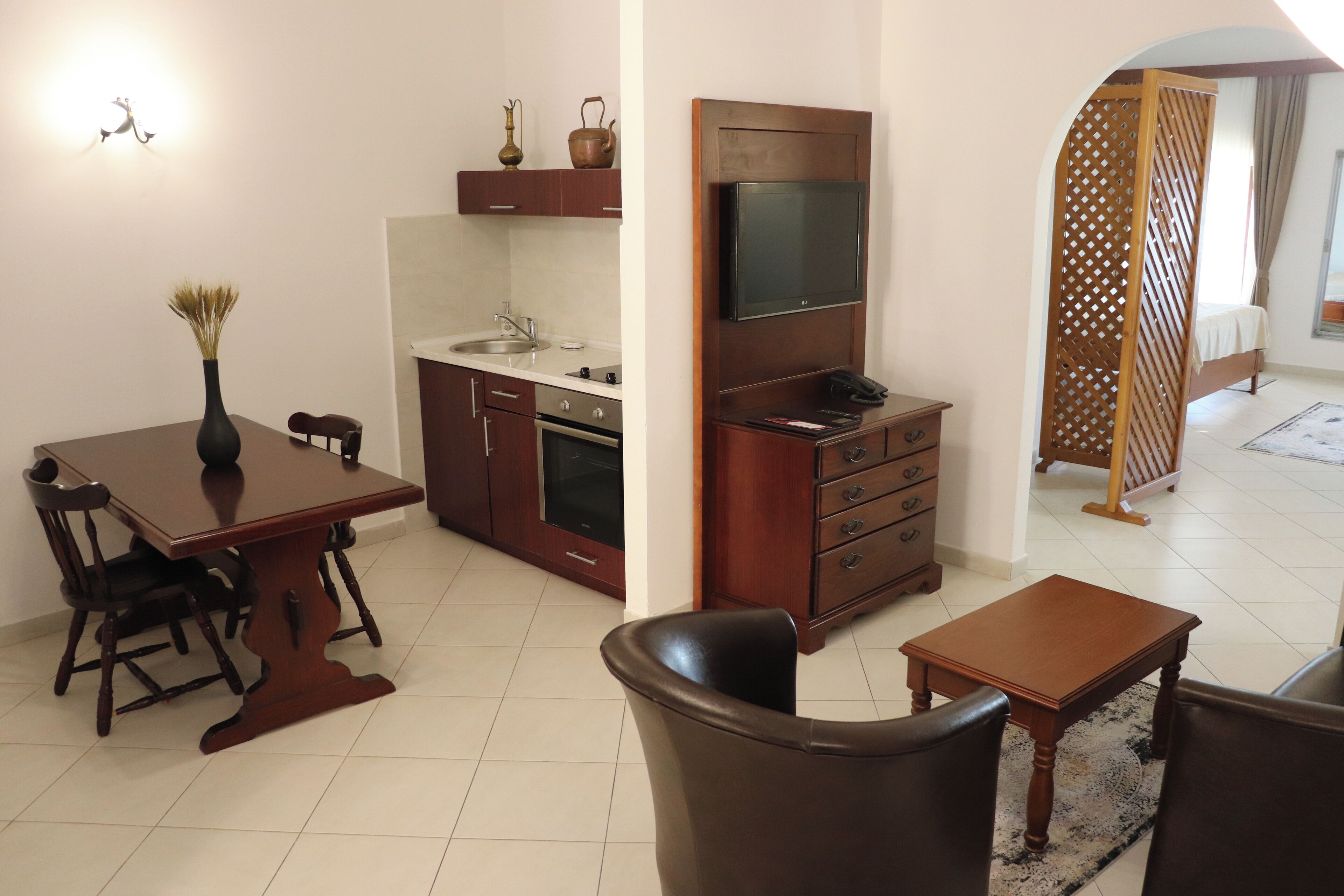A compact living space with a dining table, chairs, a kitchen area with a sink and stove, a television mounted on the wall, and a wooden partition.