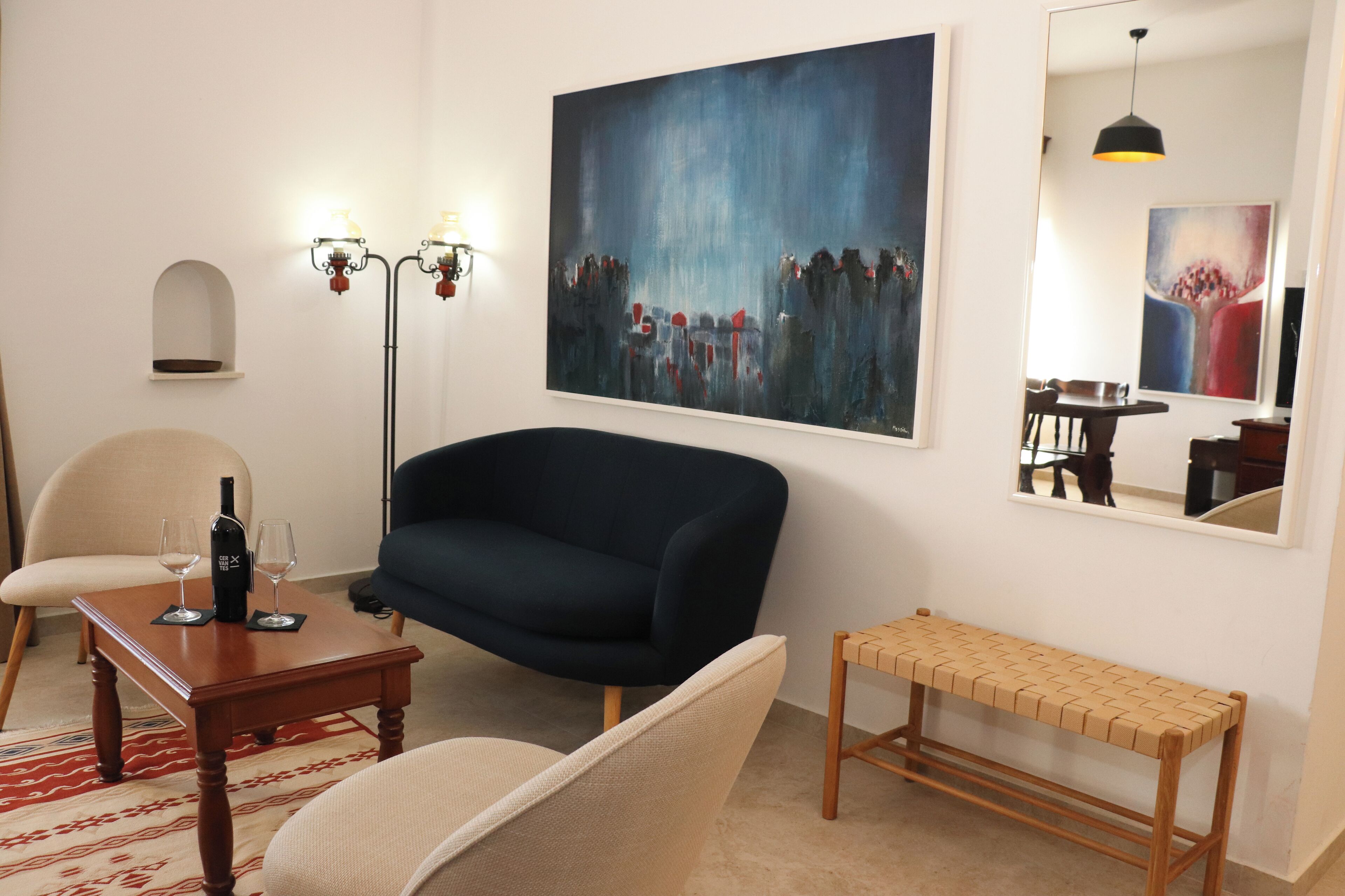 Una sala de estar con un sofá azul oscuro, una mesa de centro de madera con copas de vino, un sillón color beige, un banco tejido y un cuadro abstracto grande en la pared.