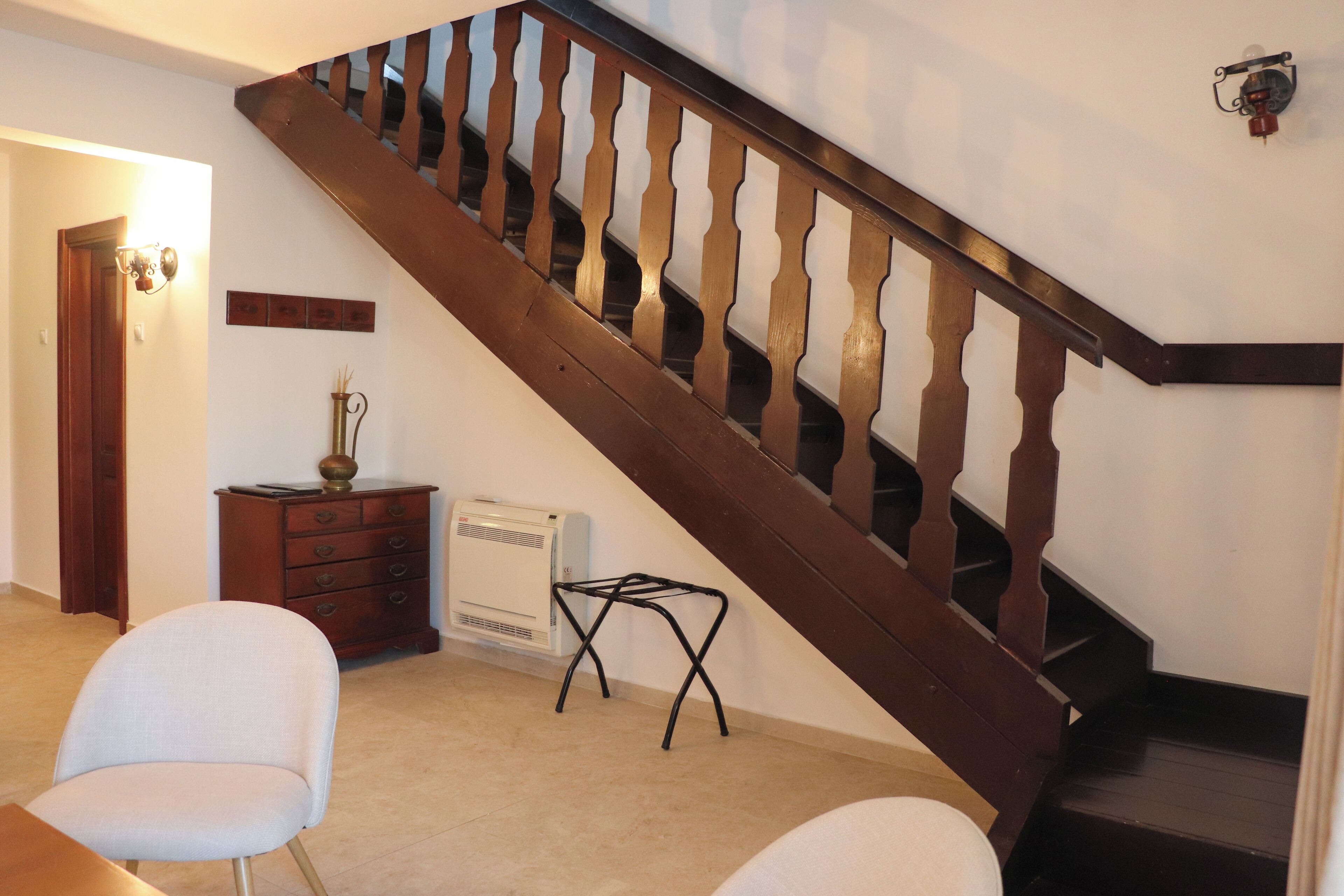 Una escalera con balaustres de madera, una mesita con un florero y una silla en una habitación con una puerta y un aplique de pared.
