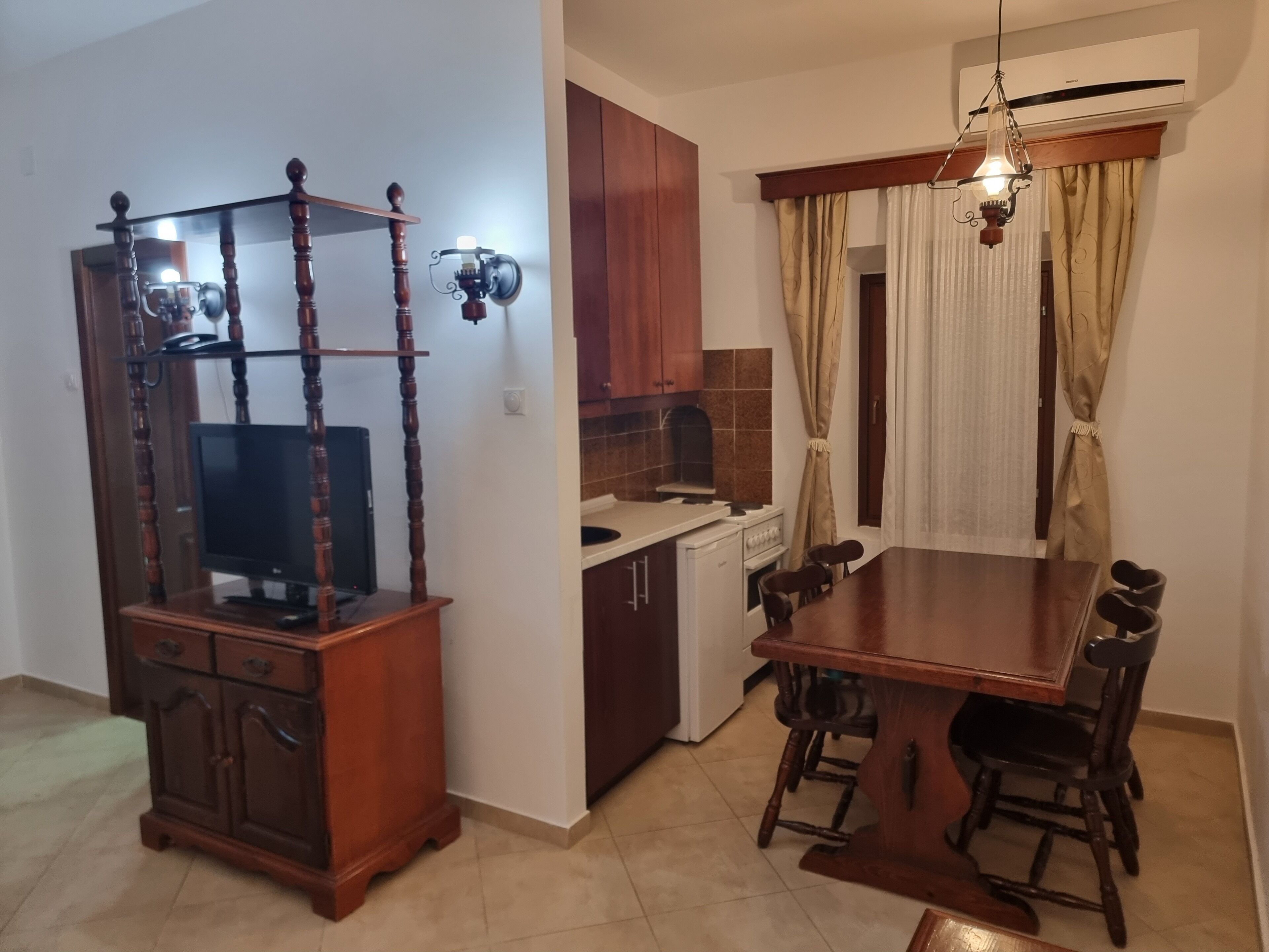 A dining area with a wooden table and chairs, a television on a wooden stand, a kitchen with cabinets and a stove, and a ceiling fan.