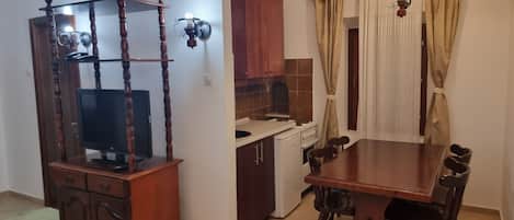 A dining area with a wooden table and chairs, a television on a wooden stand, a kitchen with cabinets and a stove, and a ceiling fan.