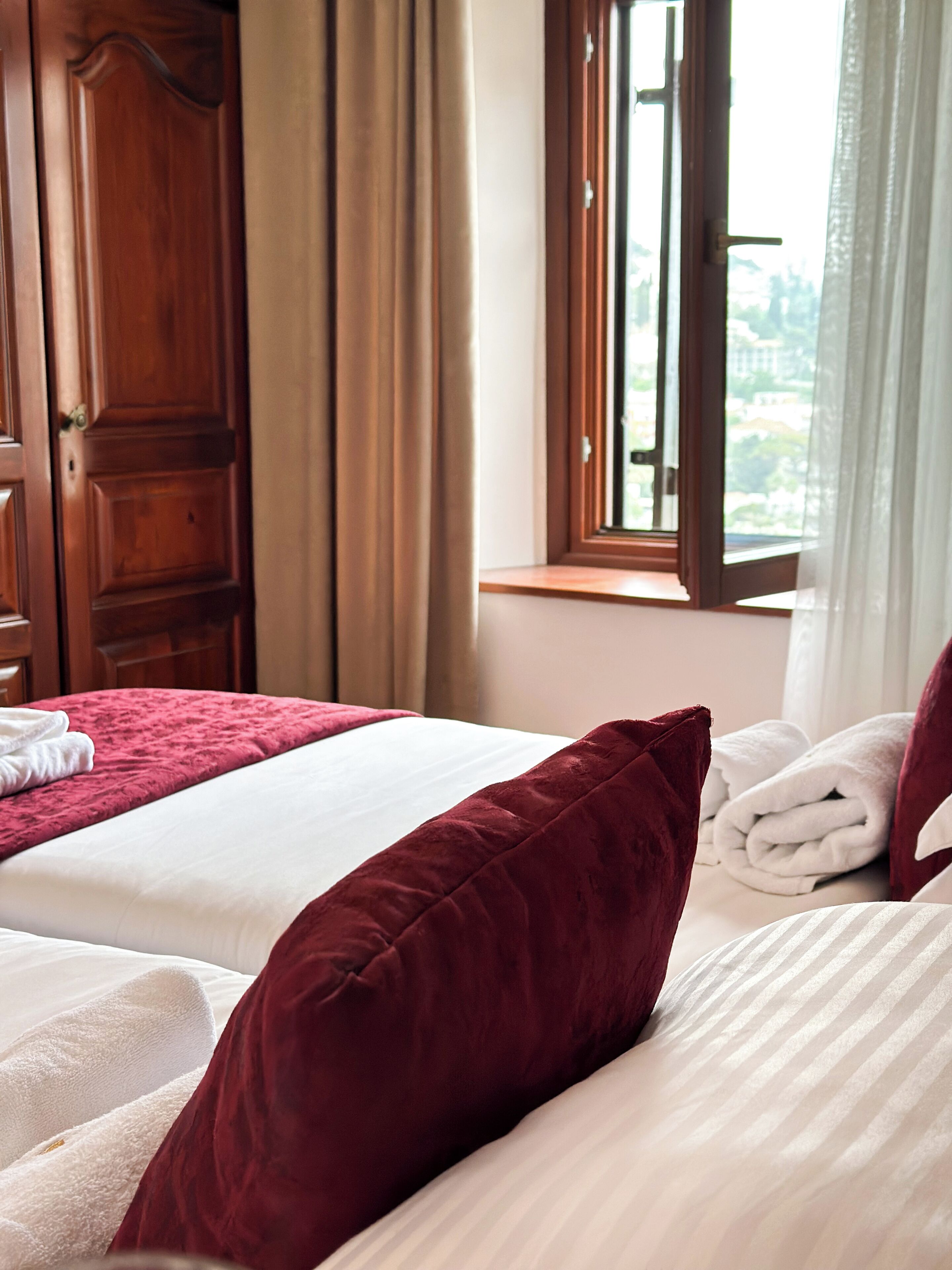 Quarto de hotel com duas camas, um guarda-roupa de madeira, uma janela com cortinas e vista para a cidade.