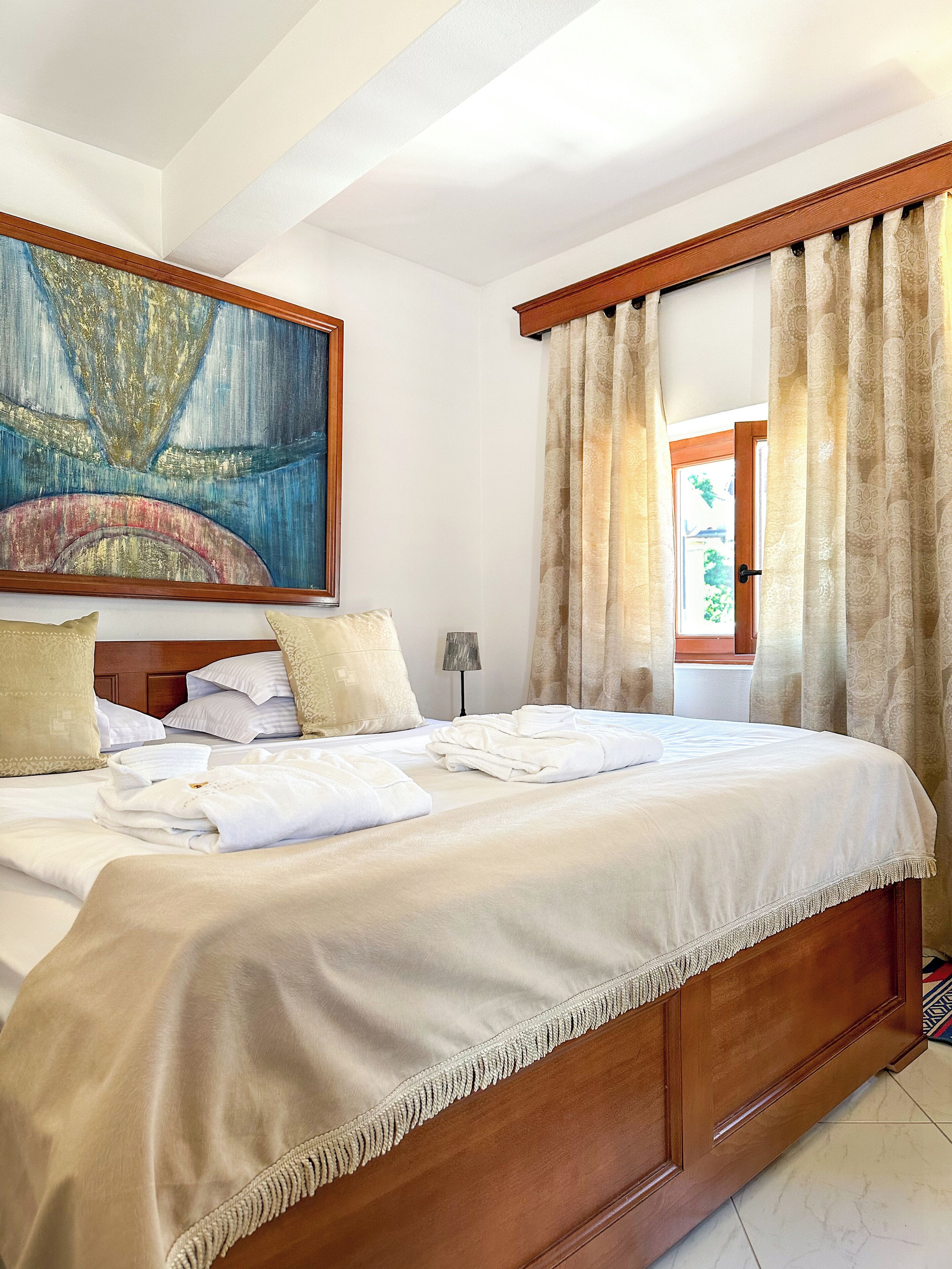 A neatly made bed with white linens, a wooden headboard, and a large abstract painting above it.
