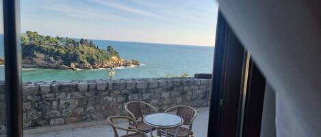 Ein Balkon mit Korbstühlen und einem Glastisch mit Blick auf die Küste, einschließlich einer Steinmauer und einem Lampenpfosten.