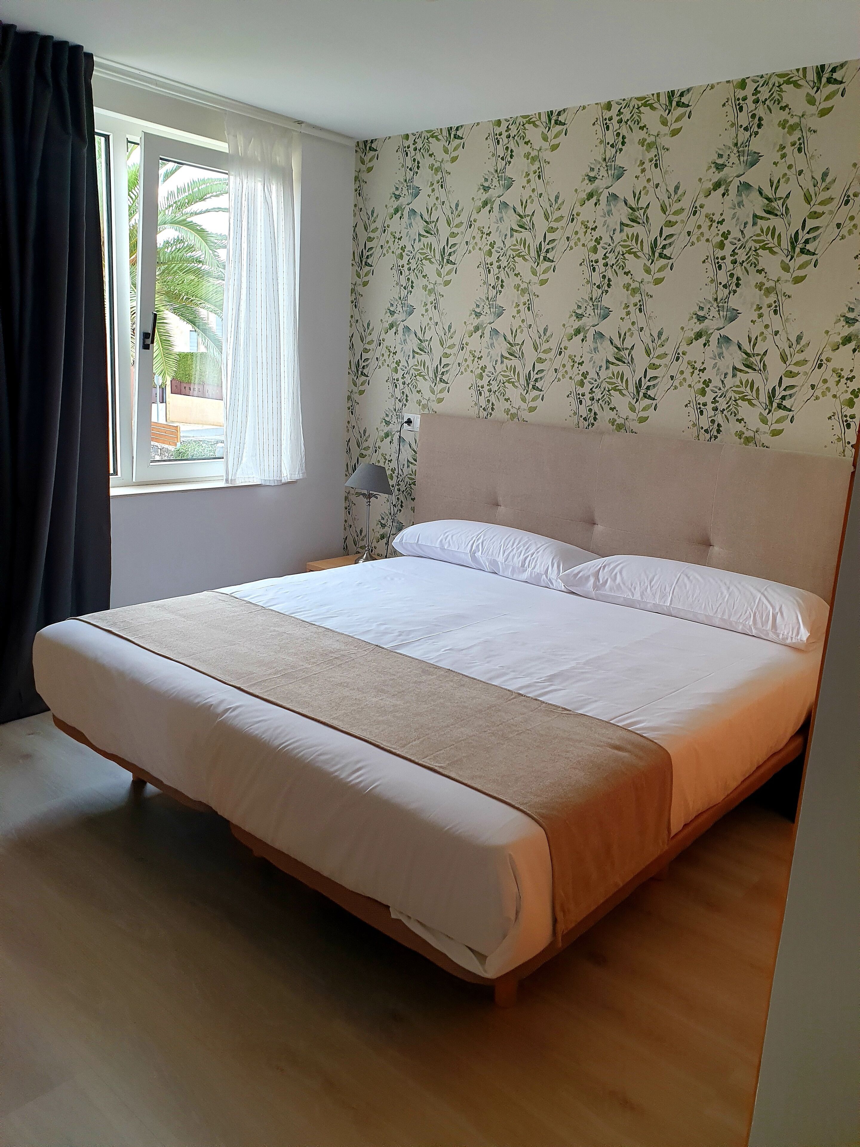 Una cama bien tendida con sábanas blancas en una habitación con papel tapiz floral, una ventana con cortinas y una mesita de noche con una lámpara.