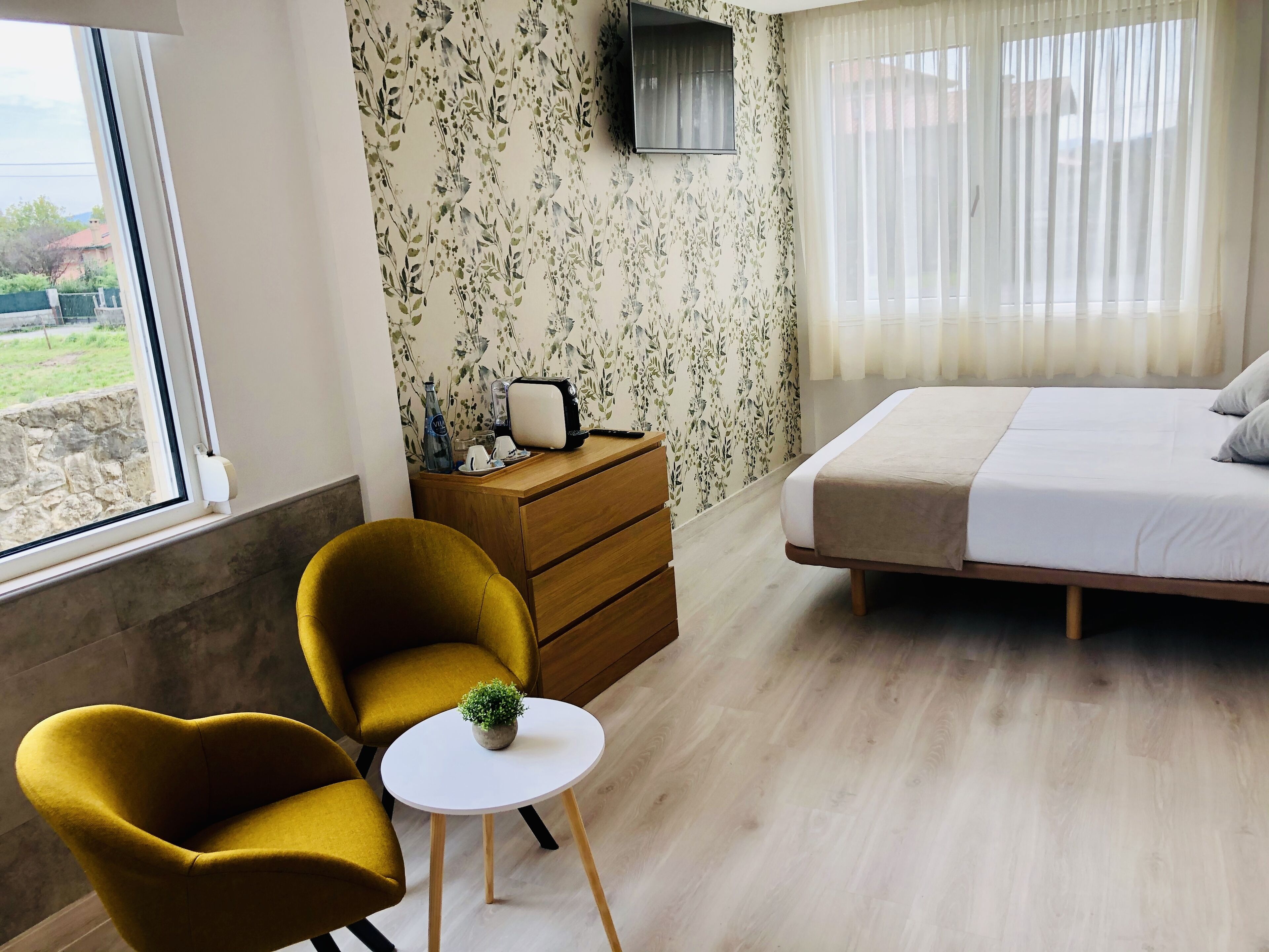 A hotel room with a bed, a wooden dresser, a small round table with a plant, two mustard-colored chairs, and a window with a view of greenery outside.