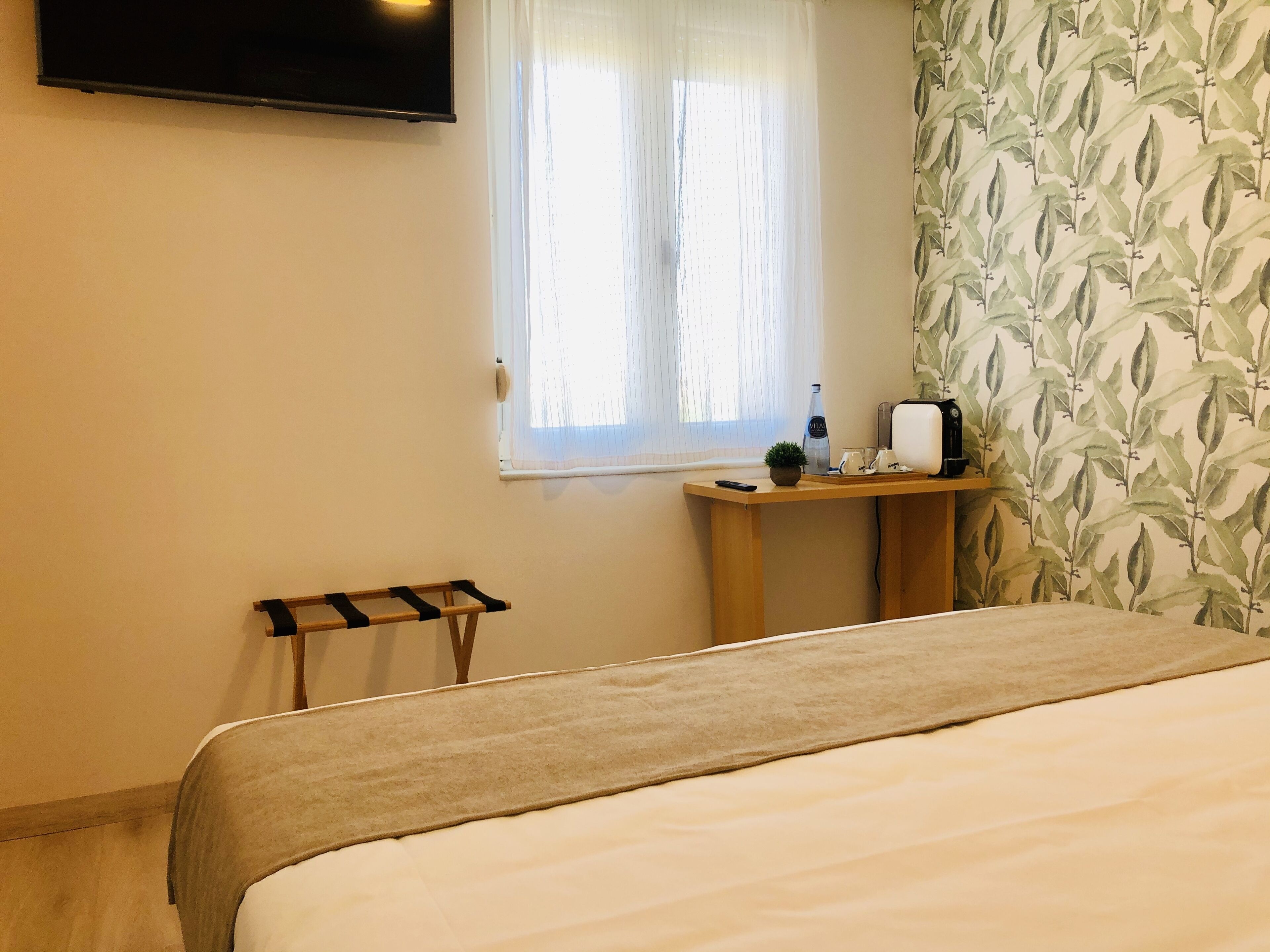 A hotel room with a bed, a TV mounted on the wall, a window with sheer curtains, a small wooden table with a remote control, a bottle, and a plant.