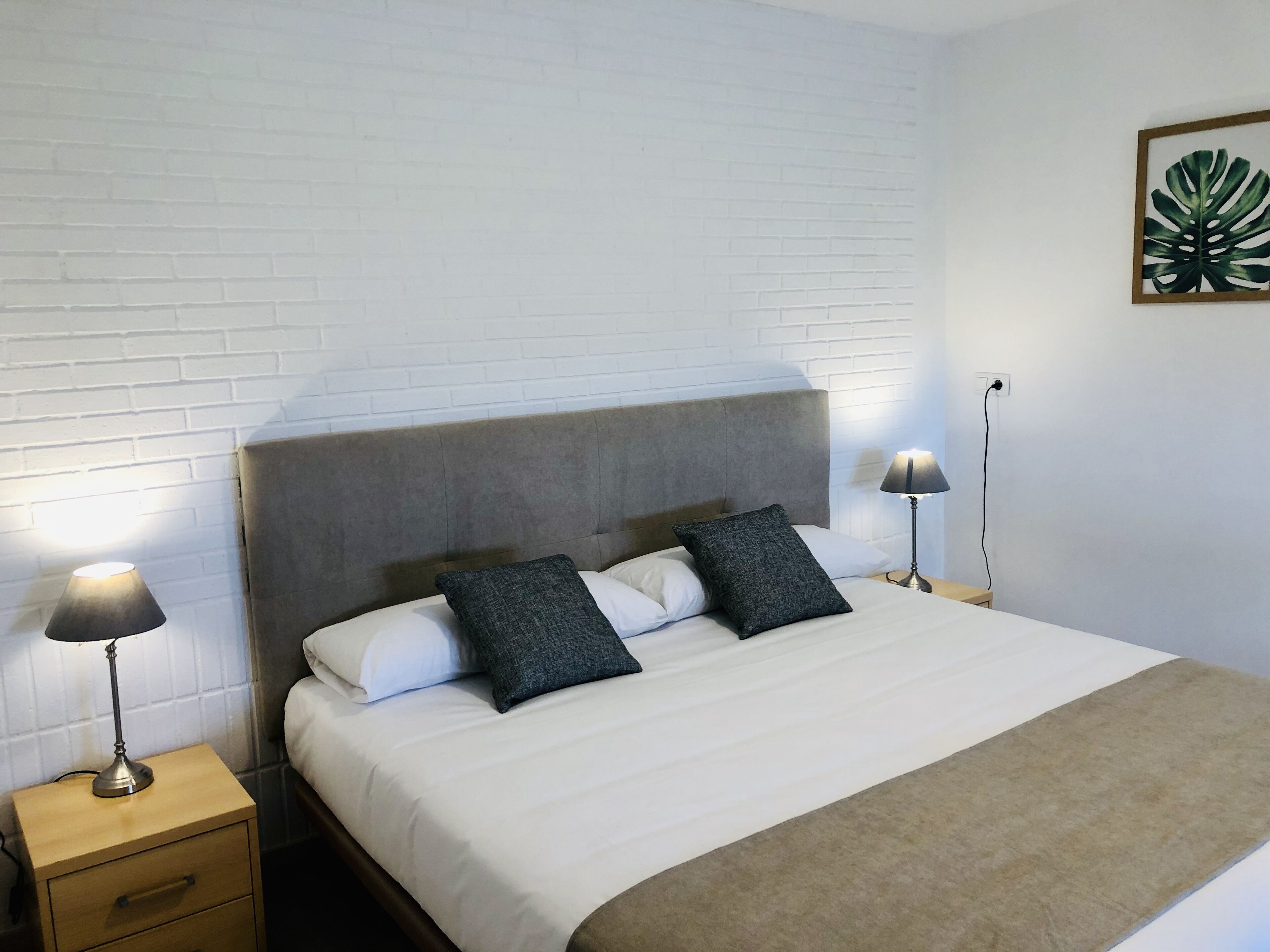 A neatly made bed with a headboard, two bedside lamps, a wooden nightstand, and a framed plant on the wall.