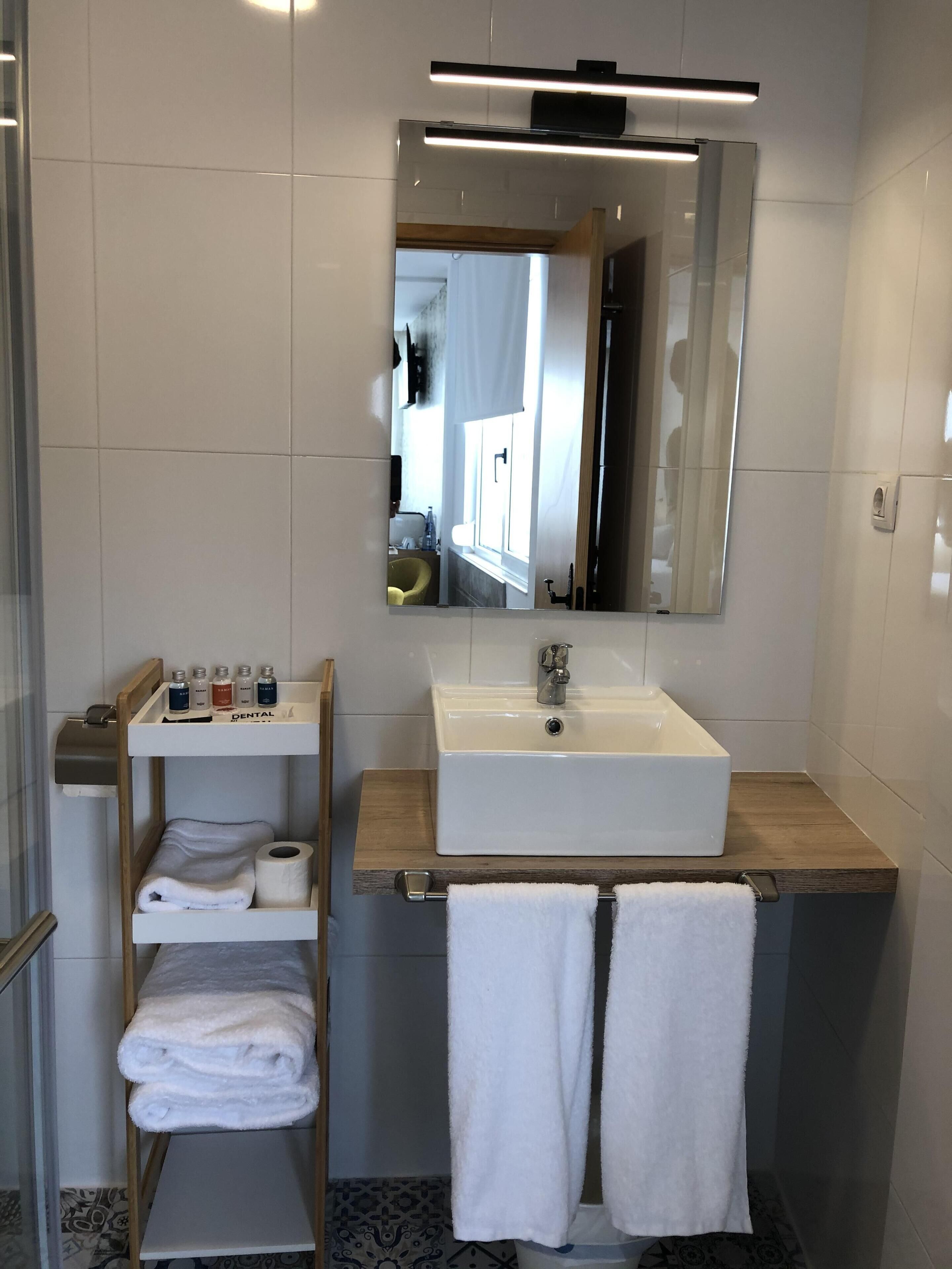 Un baño moderno con un lavabo blanco, espejo y estantes con artículos de aseo y toallas.