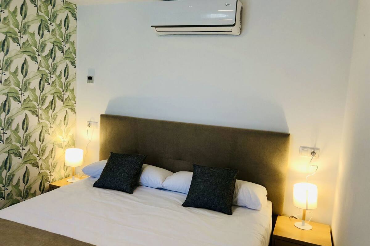 Ett hotellrum med en säng, två sänglampor, en väggmonterad luftkonditioneringsanläggning och en tapet med mönster.