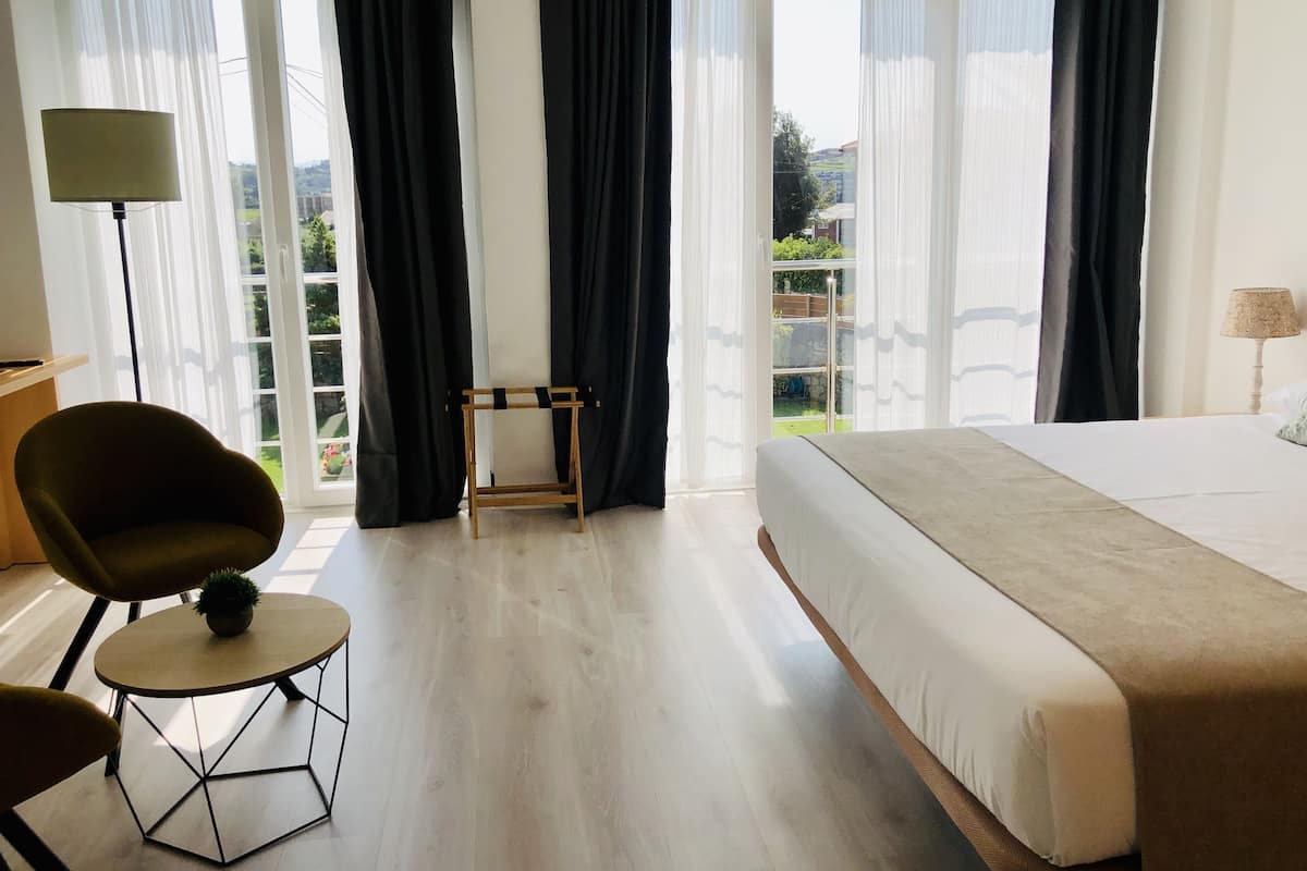 Ett hotellrum med en stor säng, en stol, ett litet bord och utsikt över omgivningarna genom stora fönster.