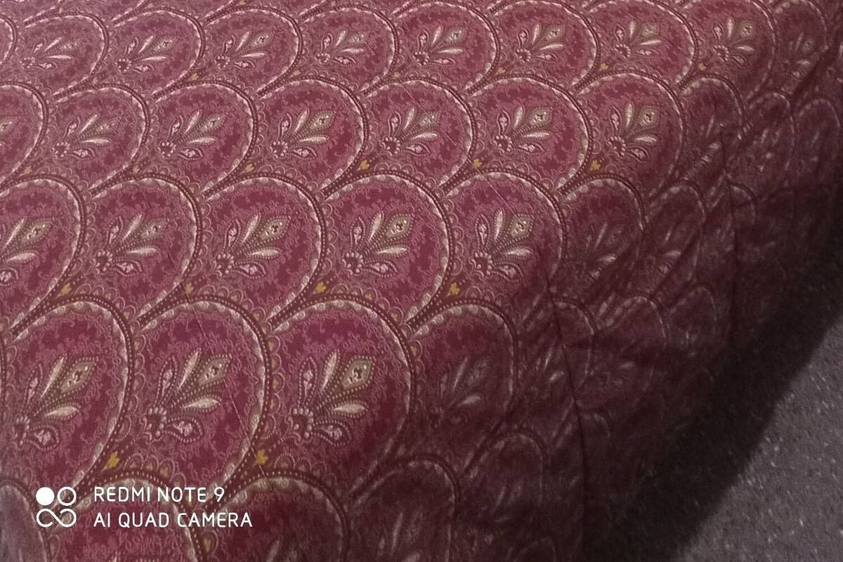 Un dormitorio con una colcha roja estampada, una silla a cuadros, un radiador y una cortina con rayas verticales.