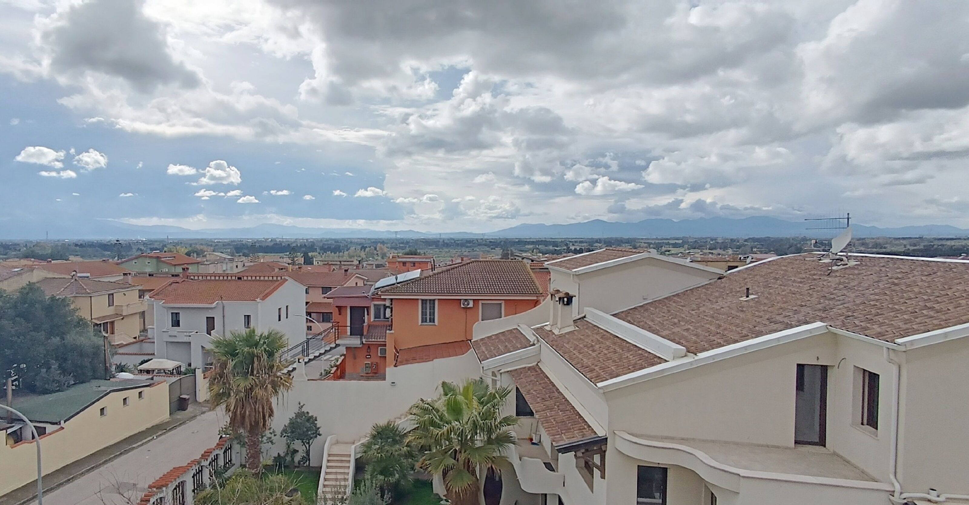 Zona residencial con casas, una calle y una cadena montañosa distante bajo un cielo nublado.