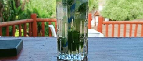 Um copo de vidro transparente com canudo e folhas de hortelã sobre uma mesa de madeira, com vista para vegetação e colinas ao fundo.