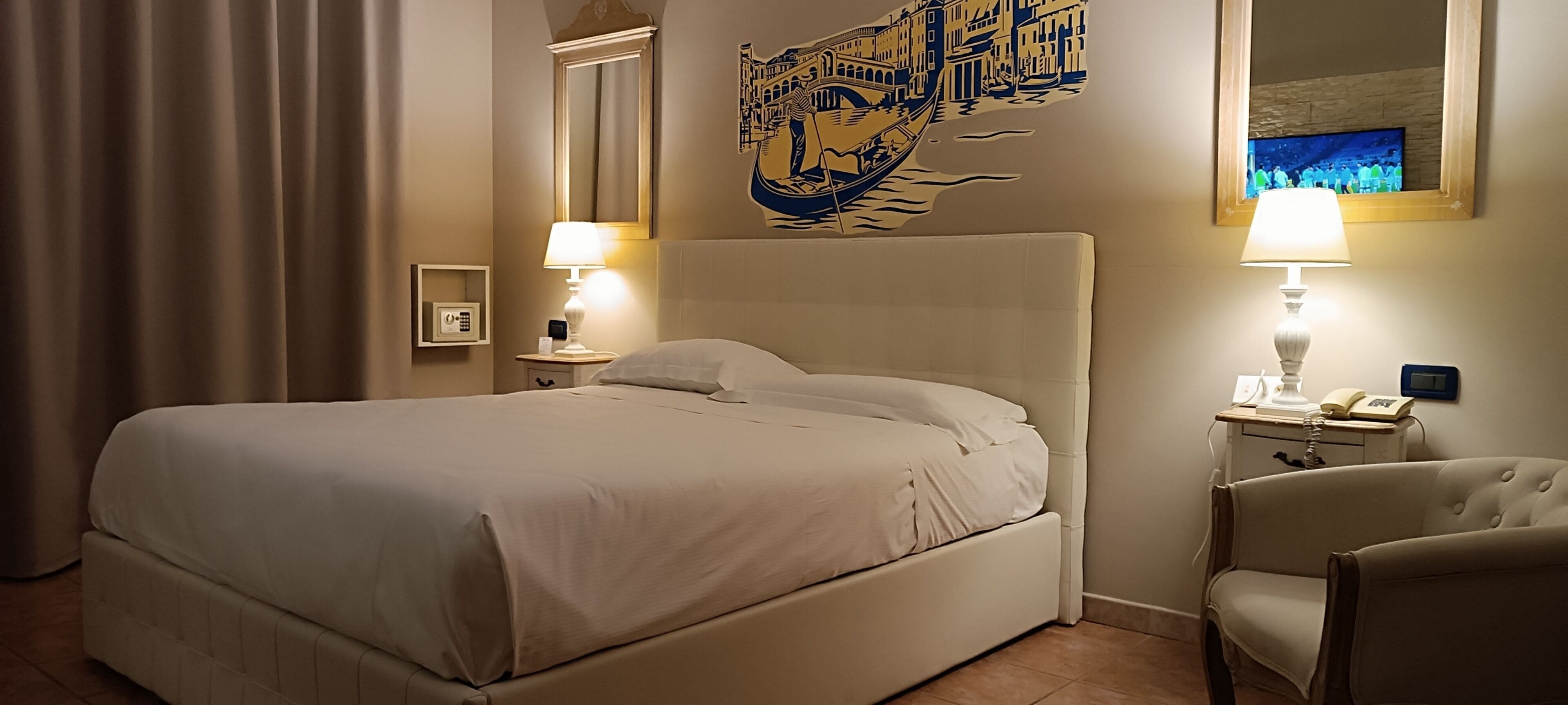Habitación de hotel con una cama grande, dos lámparas de noche, una silla, una mesita de noche con un teléfono y un cuadro de un paisaje urbano veneciano.
