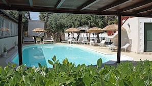 En swimmingpool med liggestole og parasoller, omgivet af grøn vegetation og et overdækket område.