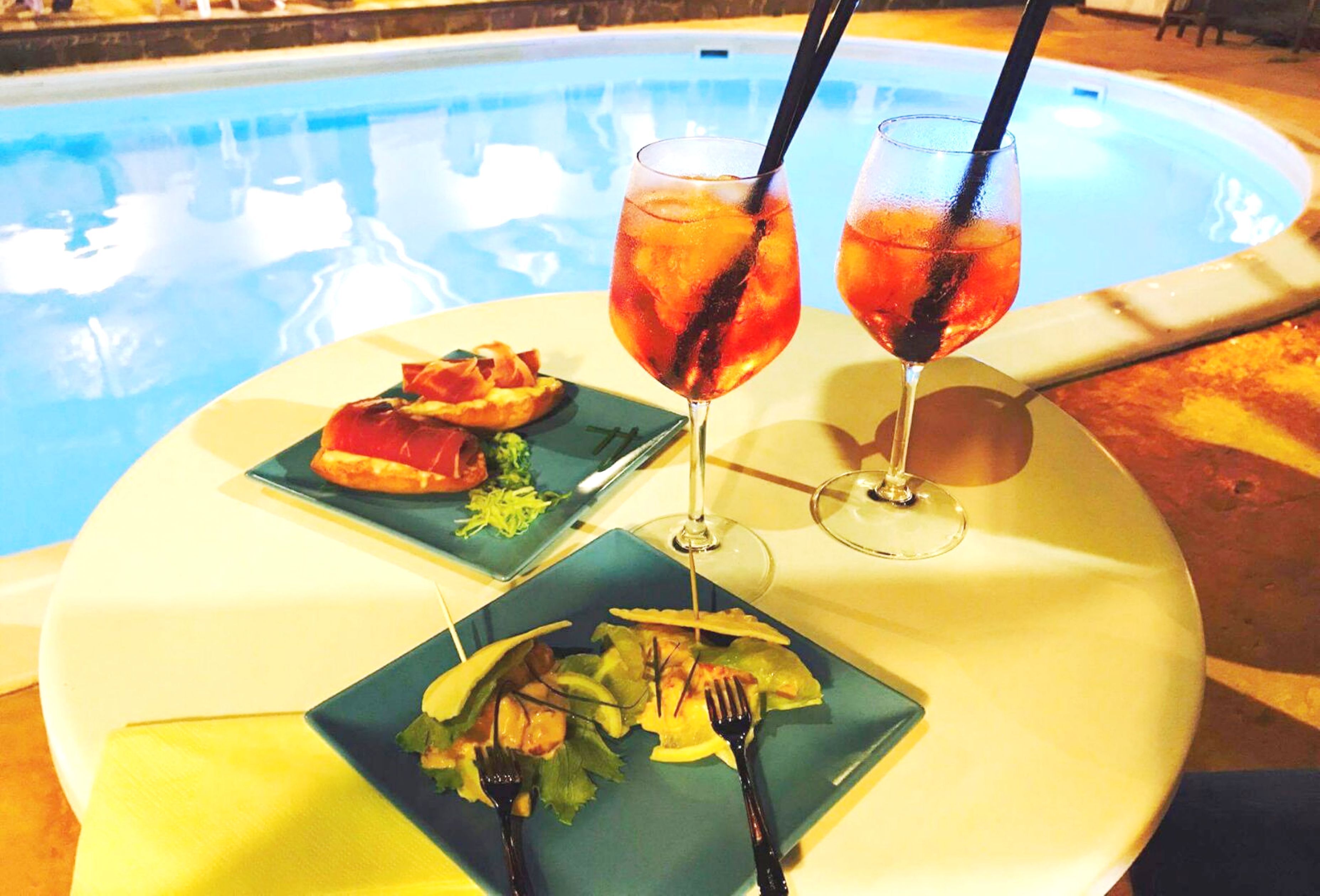 A dining table with two plates of food and two glasses of iced drinks, set by a poolside.