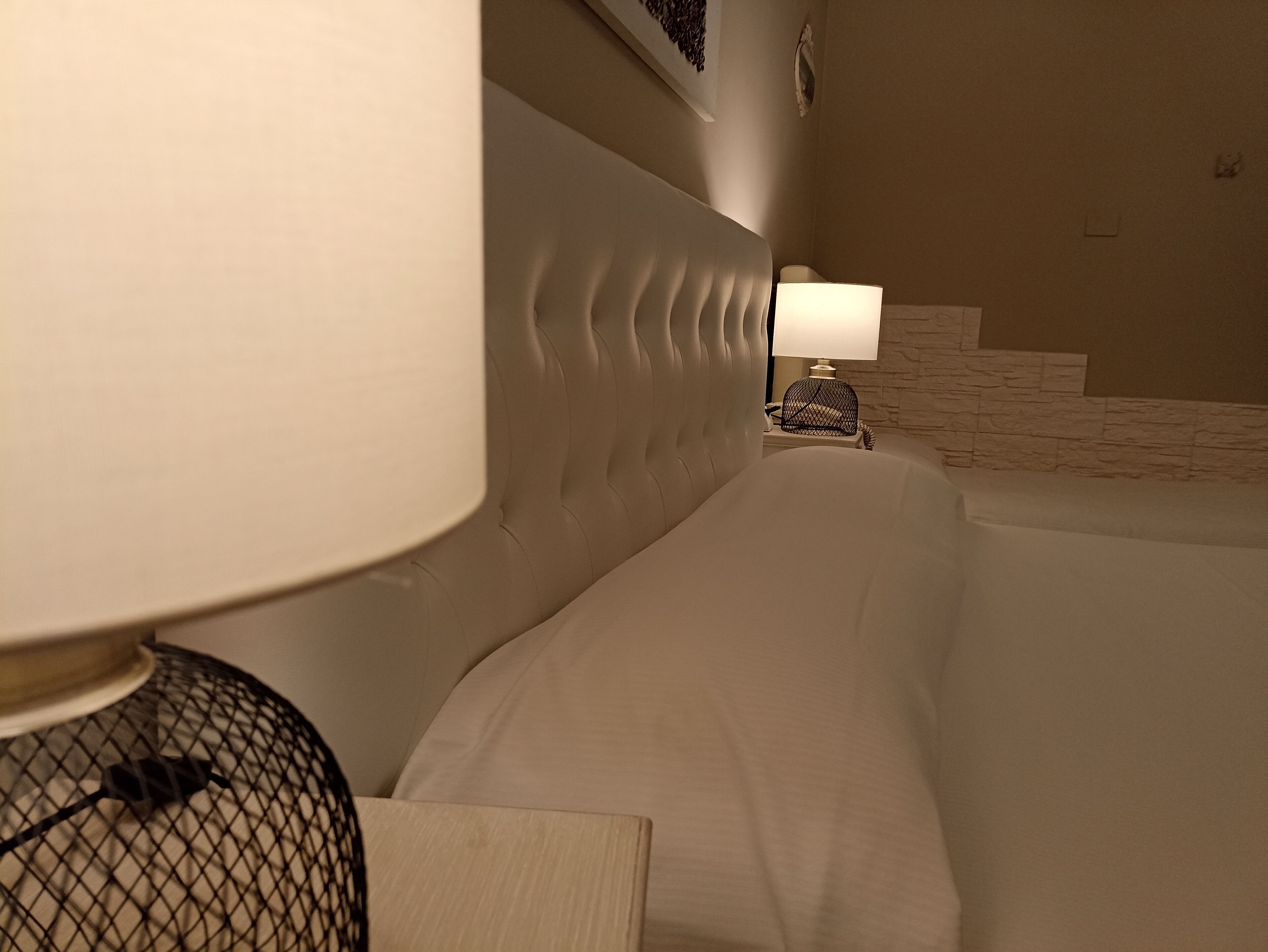 A hotel room with a large bed, a headboard, a nightstand with a lamp, and a wall-mounted shelf.