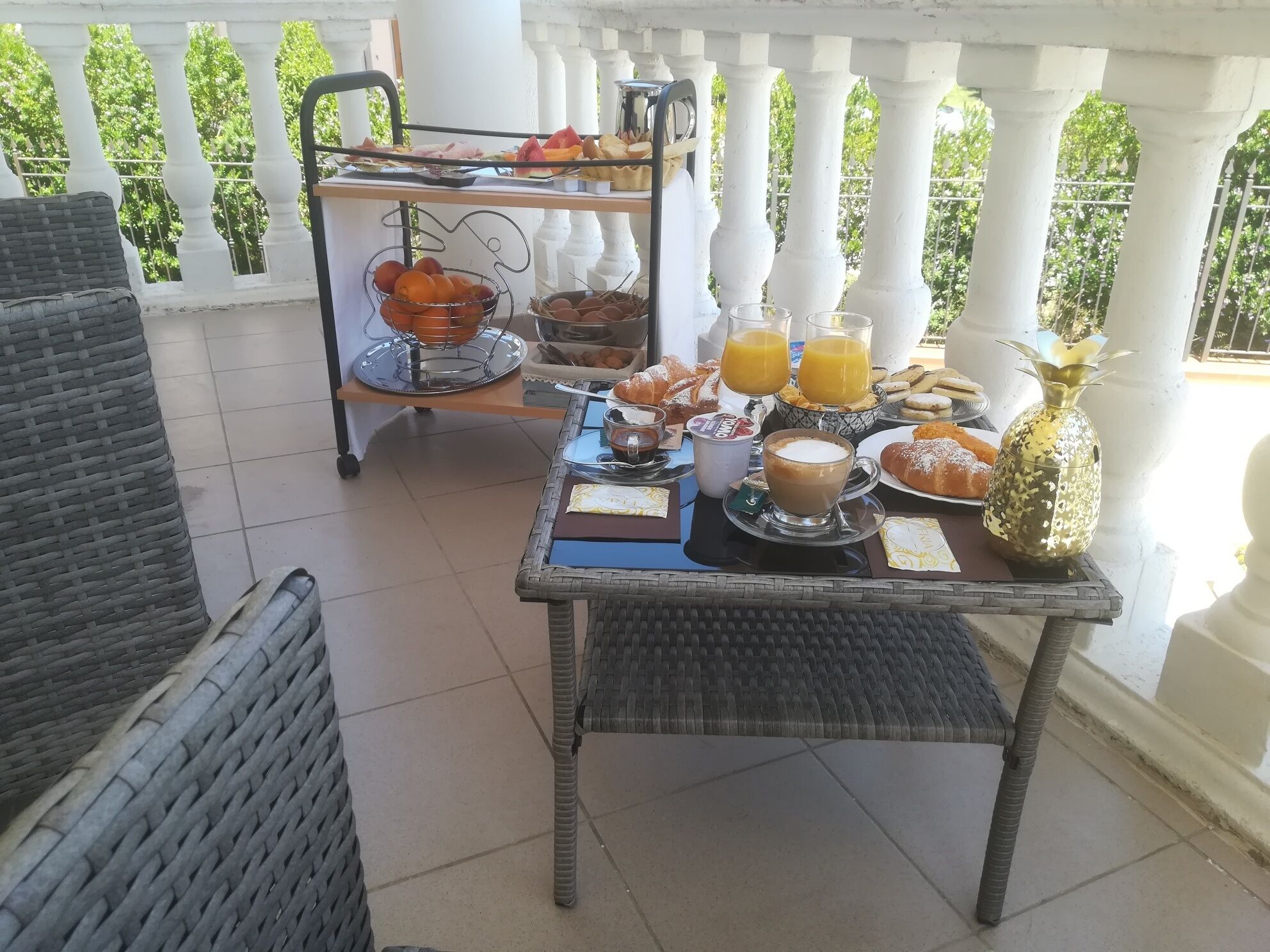 Un coin petit-déjeuner aménagé sur un balcon, avec des meubles en osier, une table garnie de nourriture et de boissons, et un chariot contenant des articles supplémentaires.