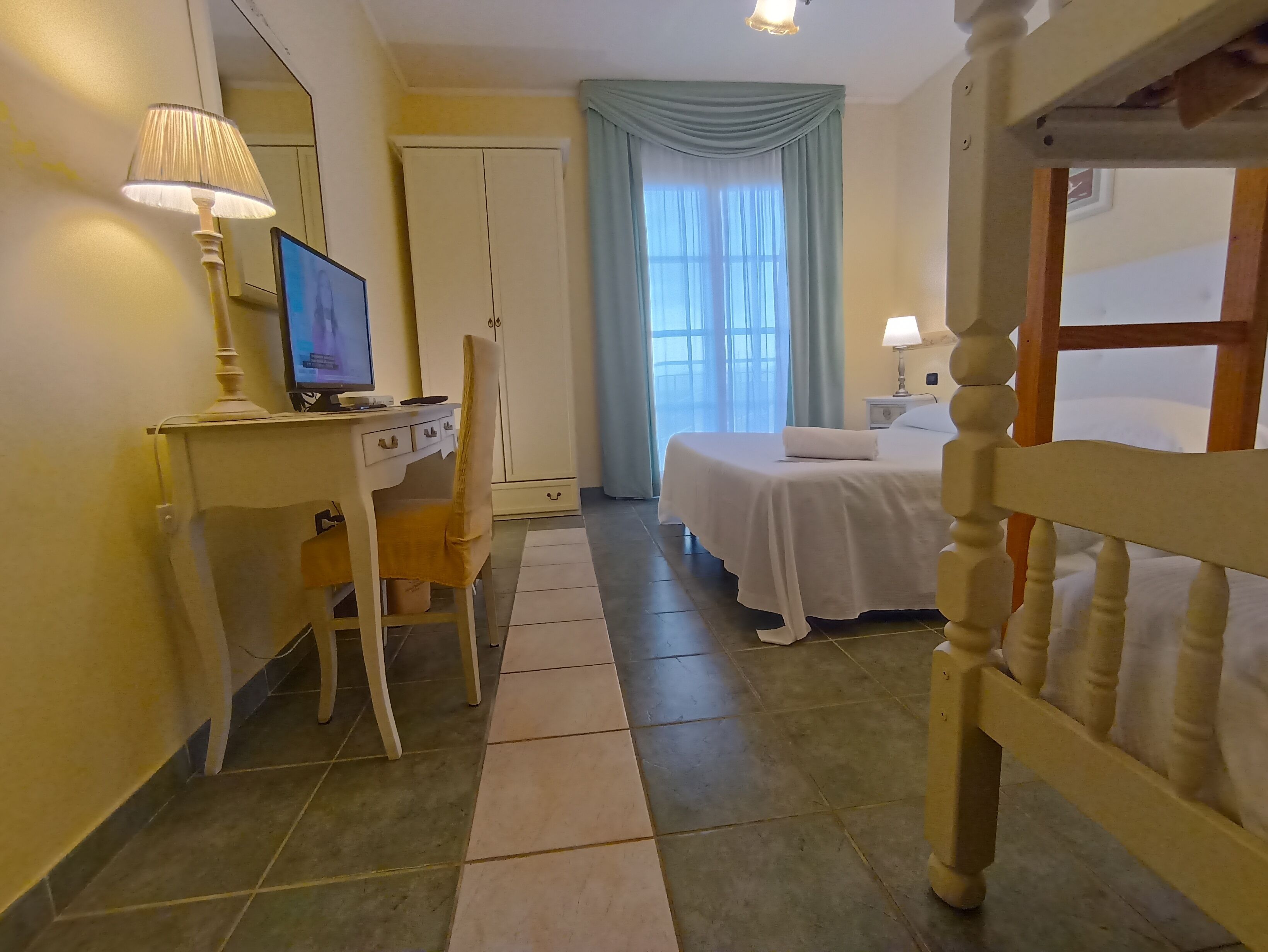 Ein Hotelzimmer mit Bett, Schreibtisch mit Computer, Stuhl, Kleiderschrank und Fenster mit Vorhängen.