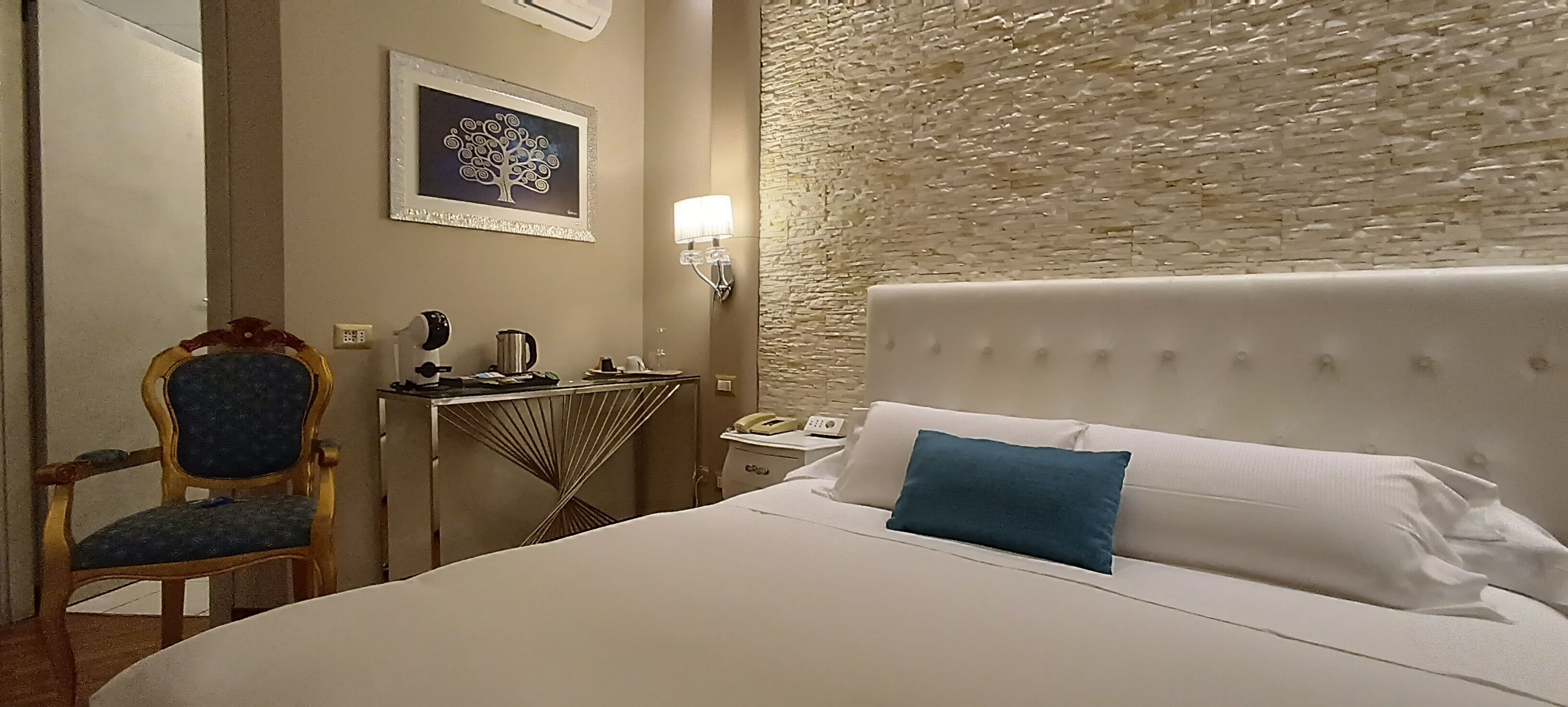 Habitación de hotel con cama, una silla, una mesita con hervidor, lámpara y tel éfono en la pared.