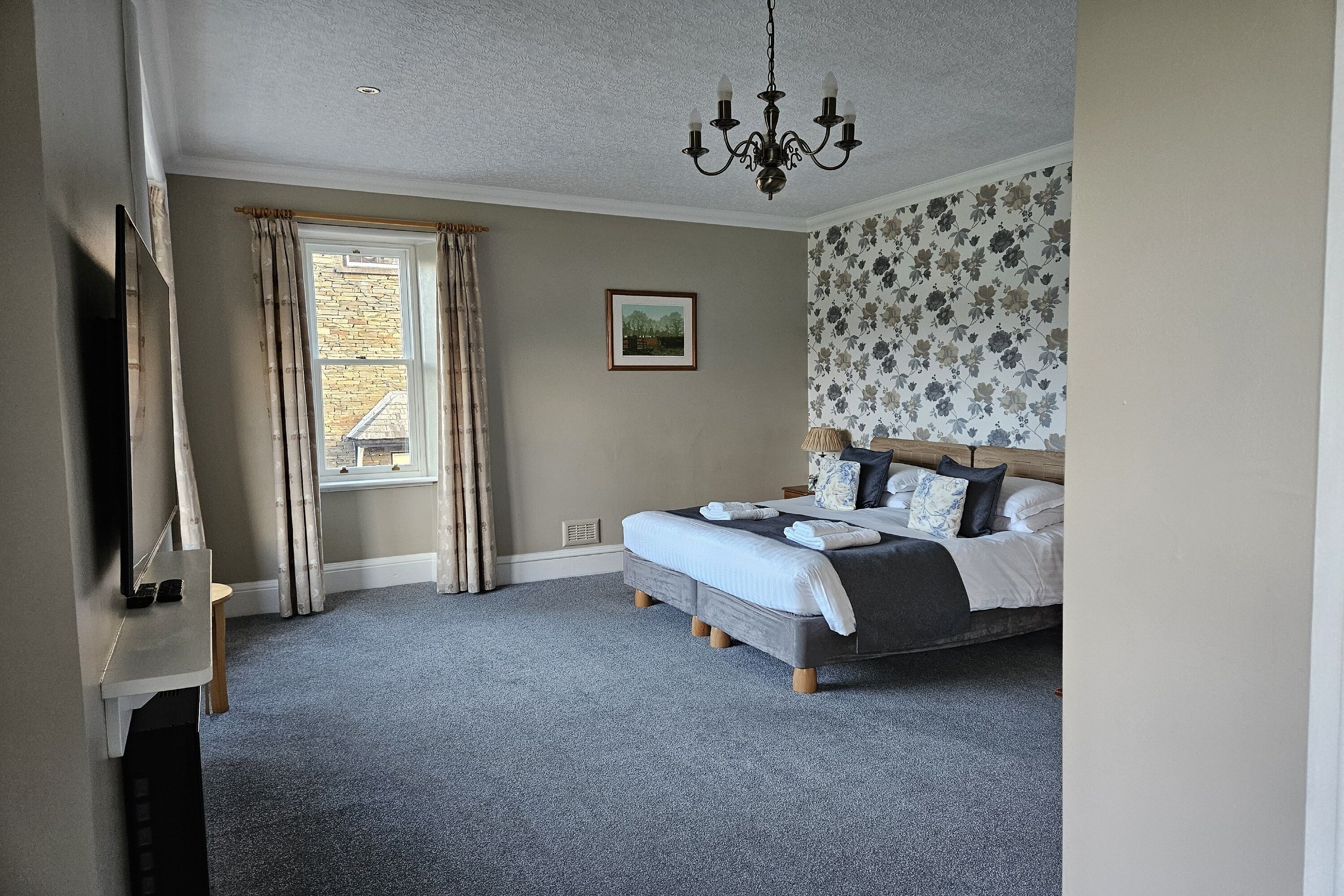 Ein Schlafzimmer mit Bett, einer Kristallleuchter, einem Fenster mit Vorhängen, einem gerahmten Bild an der Wand und einem Teppichboden.