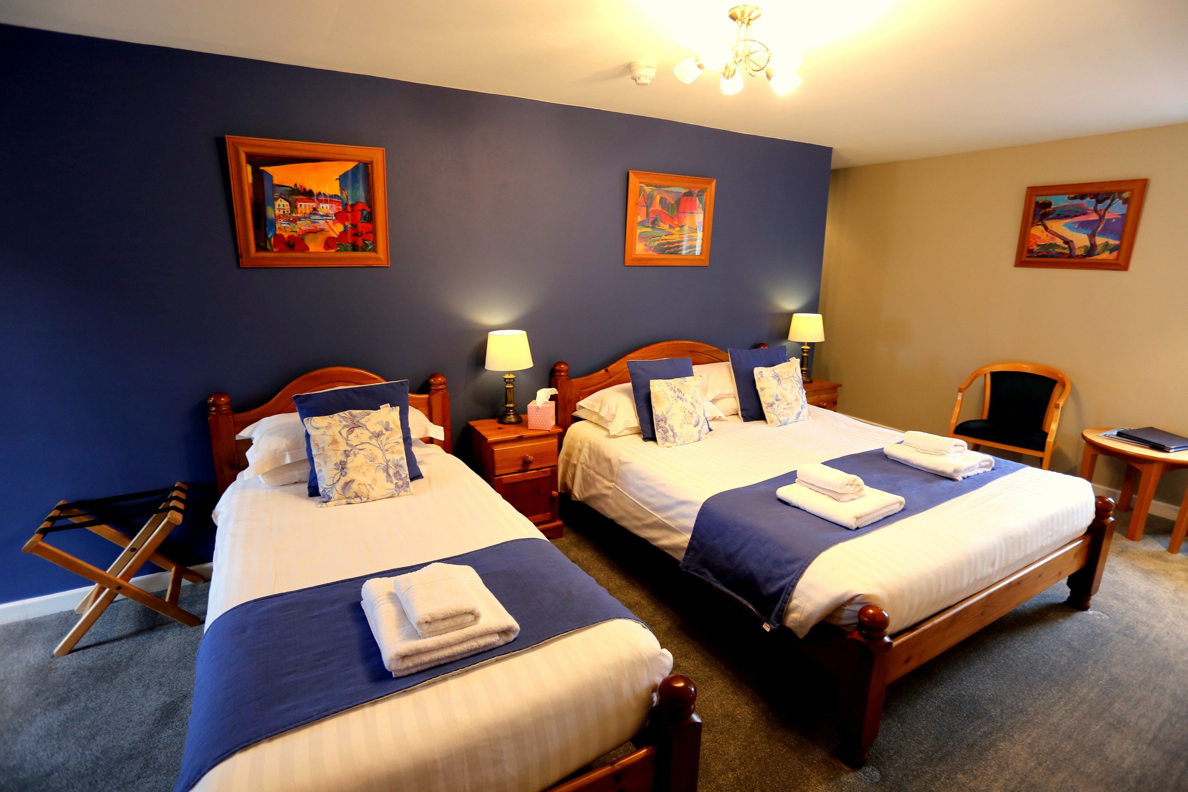 A hotel room with two beds, each with a wooden headboard, blue bedding, and white towels. There are paintings on the walls and a small table with a lamp.