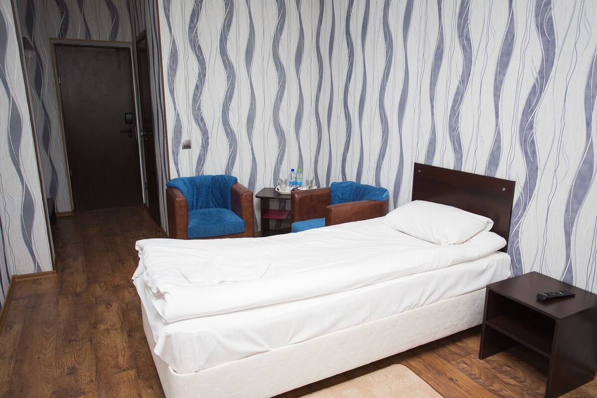 Hotellihuone, jossa on sänky, kaksi sinistä nojatuolia, pieni pöytä, jossa on vesipullo, ja yöpöytä, jossa on kaukosäädin.