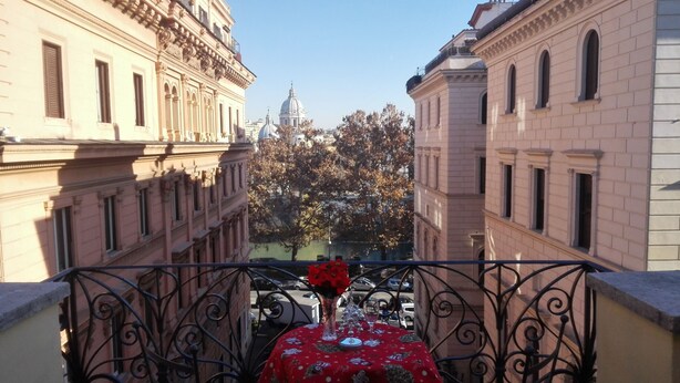 Un balcón con una mesa lista para dos personas, con vista a una calle de arquitectura clásica y un árbol con flores rojas.