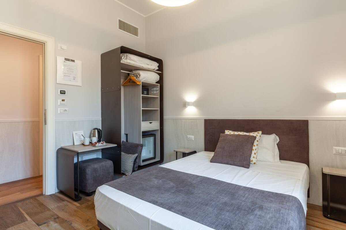 A hotel room with a bed, bedside table, wardrobe, and a desk with a chair.