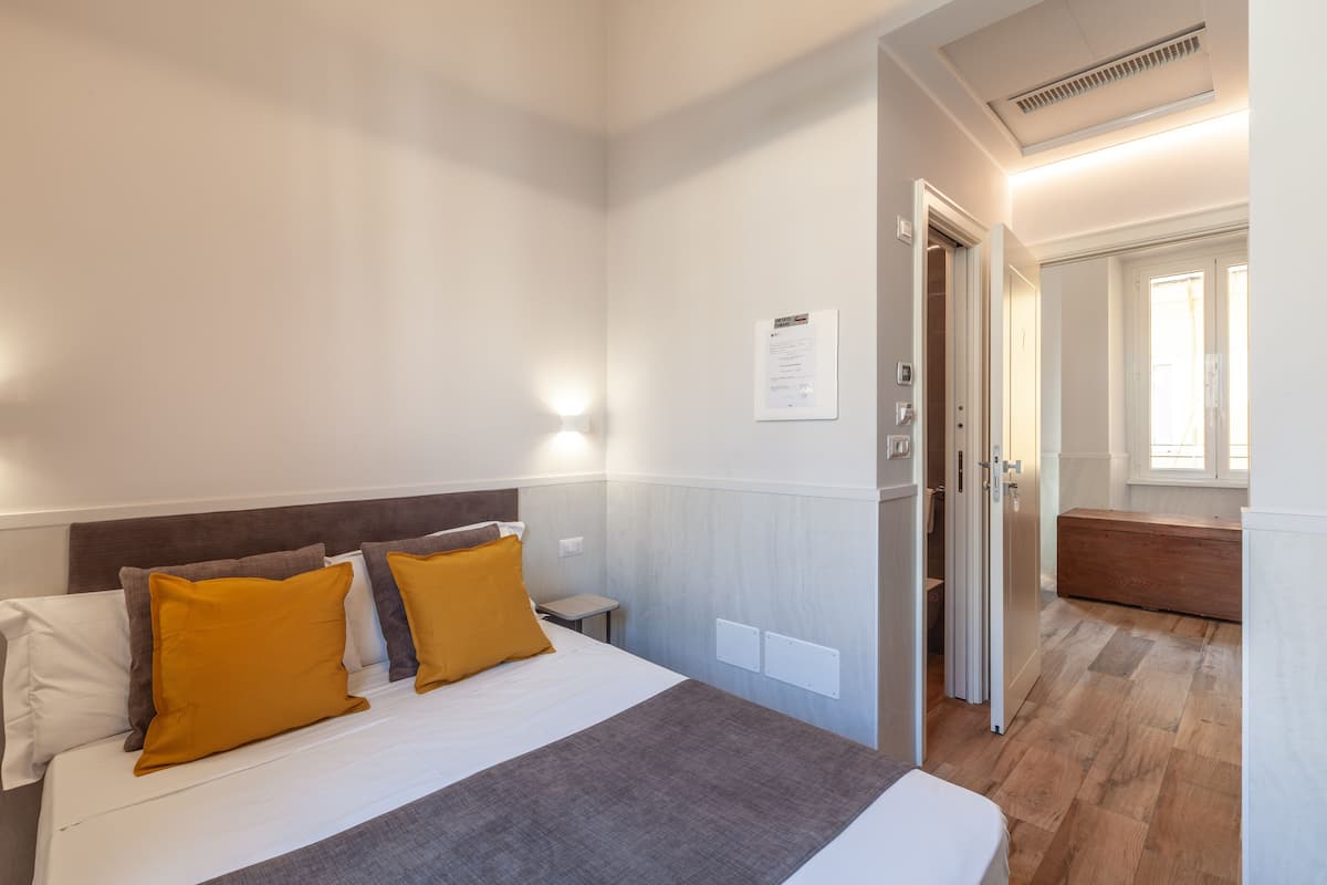 A hotel room with a bed, a nightstand, a wooden bench, and a door leading to another room.