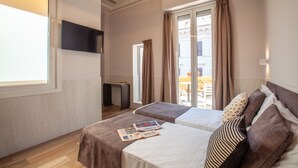 Habitación de hotel con dos camas, un televisor montado en la pared, un escritorio pequeño y una ventana con cortinas.