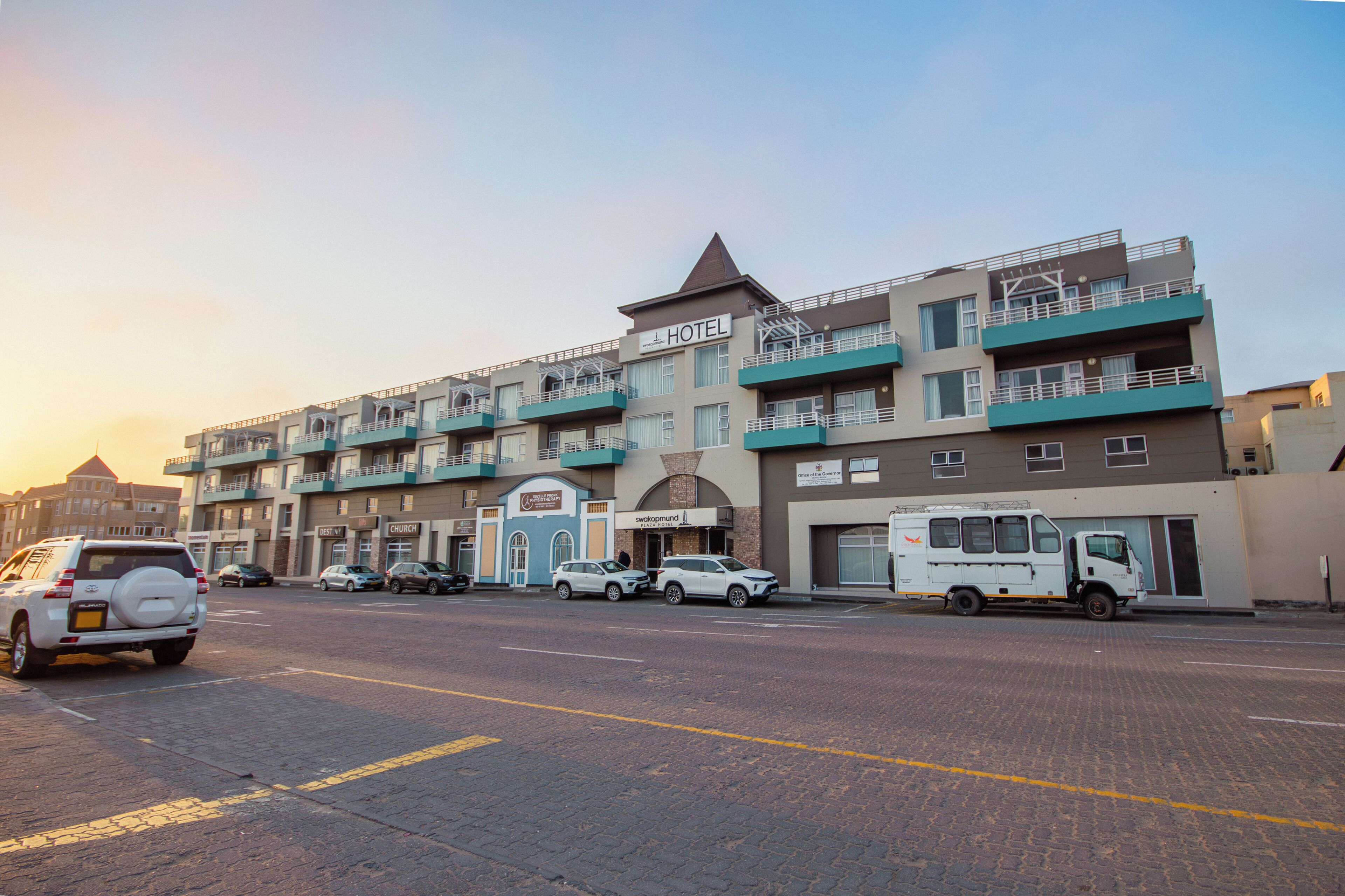 Un hotel de varios pisos con techo plano, varios balcones y un estacionamiento con varios coches.