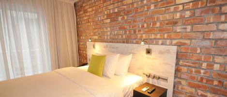 A hotel room with a brick wall, a bed with white bedding, a nightstand with a remote control, and an air conditioning unit.
