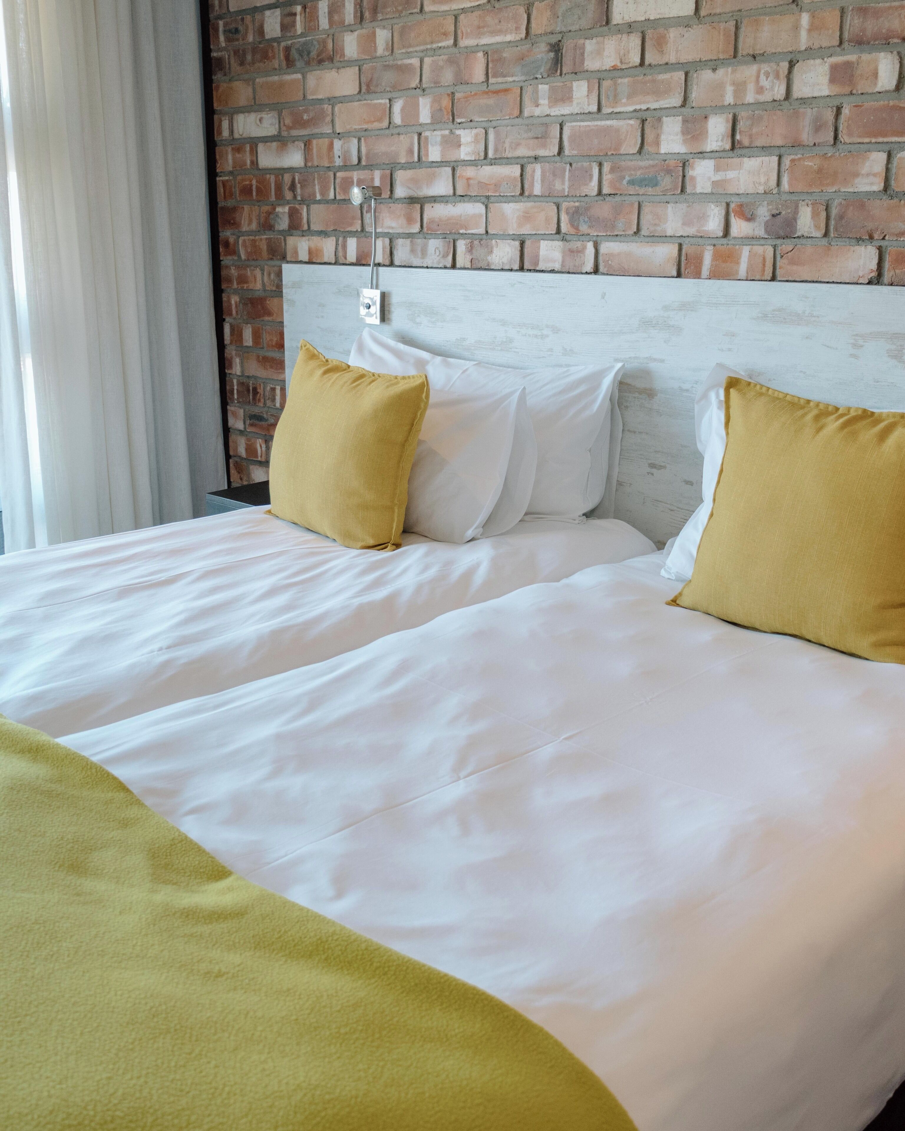 A bed with white bedding and two yellow pillows, set against a brick wall.