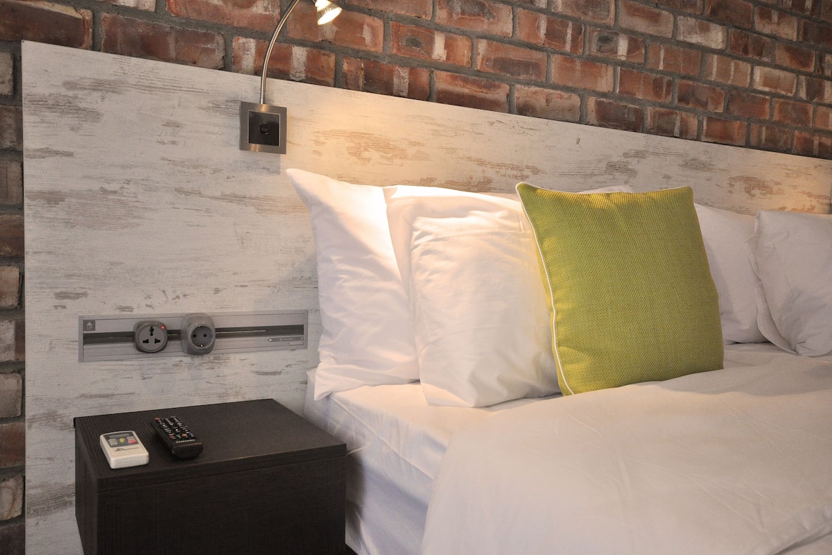 A bed with white bedding and a green pillow, a nightstand with a remote control, and a brick wall with a power outlet.