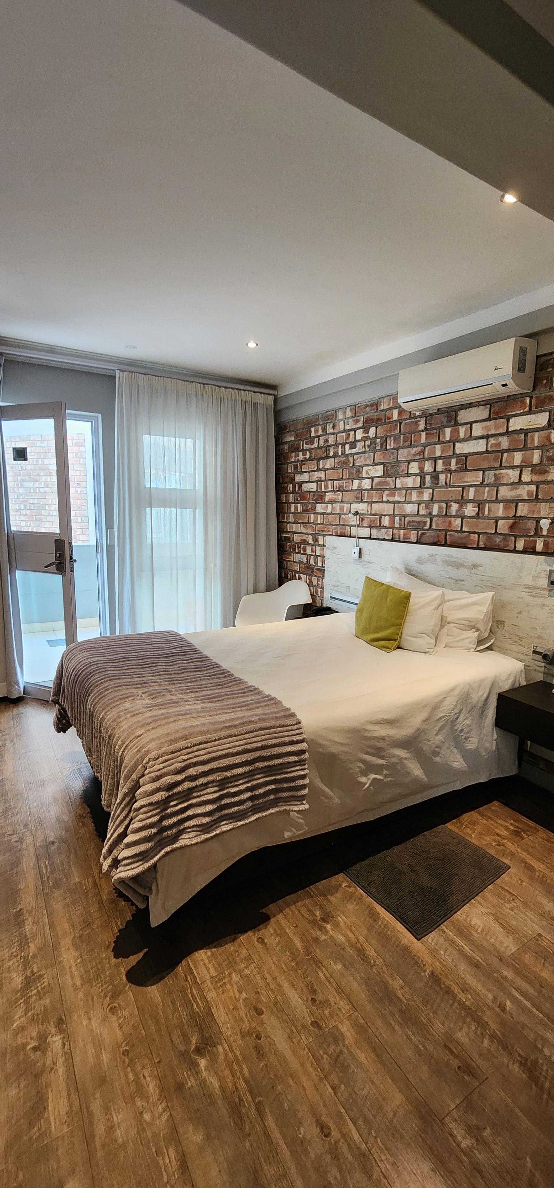 A modern bedroom with a brick wall, a large bed with a striped blanket, a bedside table, and a window with curtains.