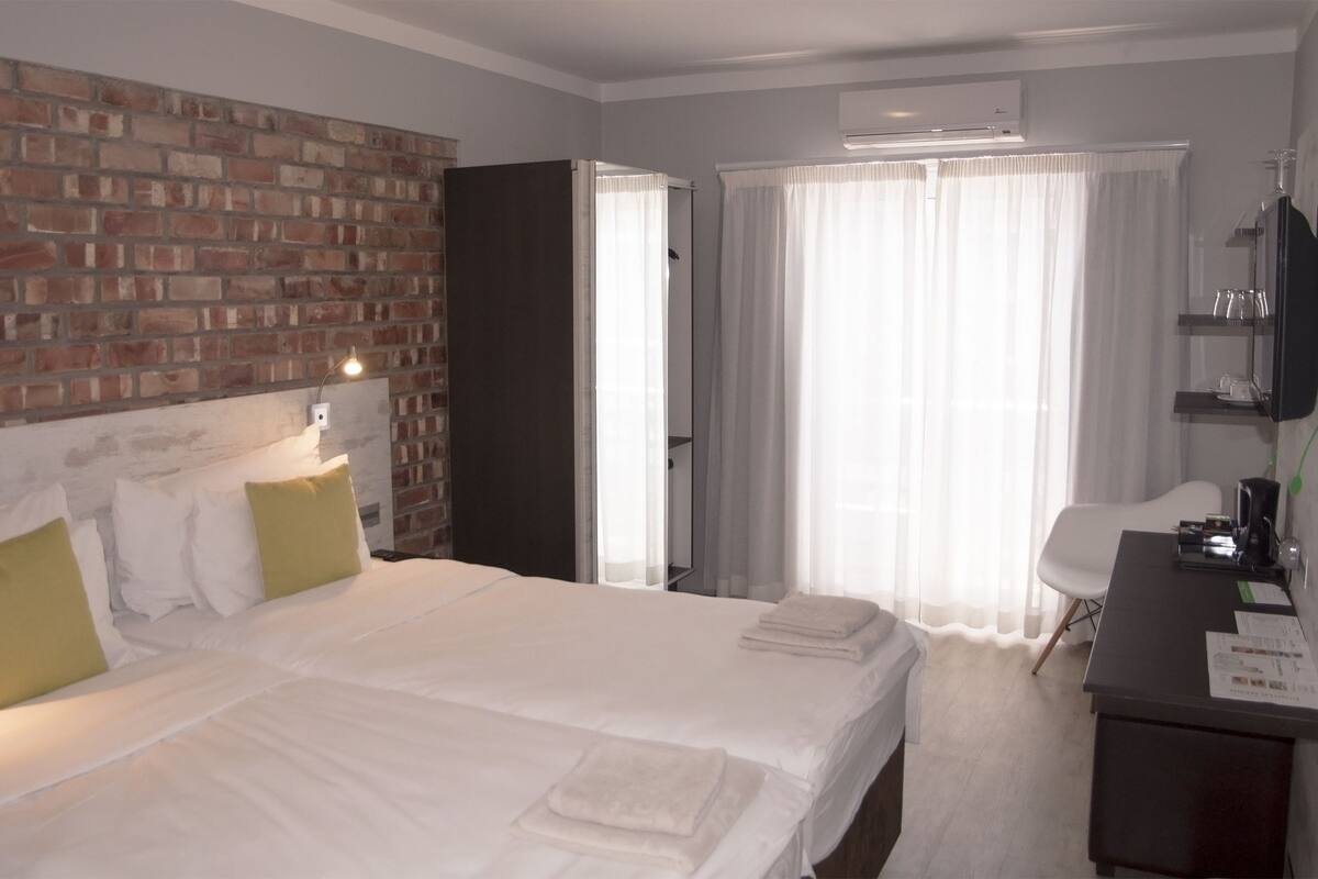 A hotel room with a brick wall, a bed with white linens, a desk with a chair, and a window with sheer curtains.