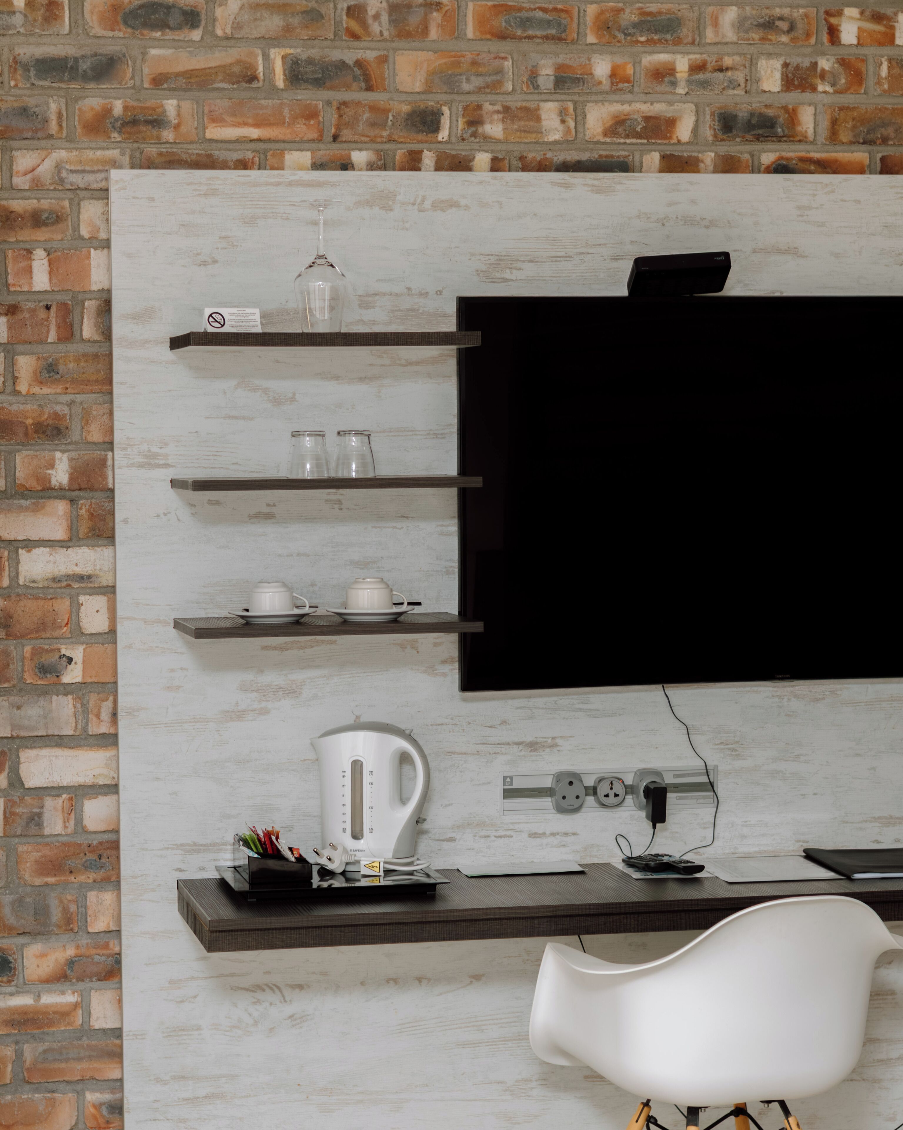 A modern kitchen with a brick wall, a flat-screen TV mounted above a wooden shelf, and a white chair.