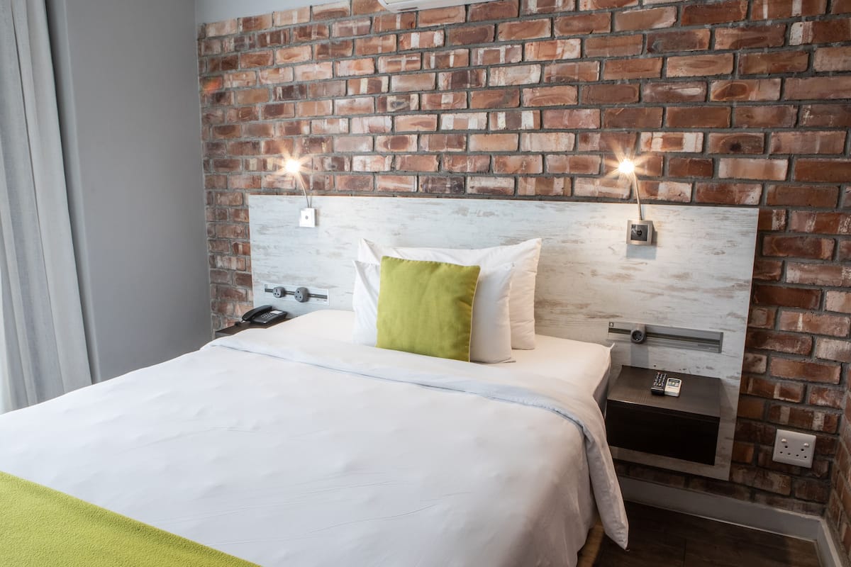 A hotel room with a brick wall, a bed with white bedding and a green pillow, a bedside table with a phone and a lamp, and a power outlet on the wall.