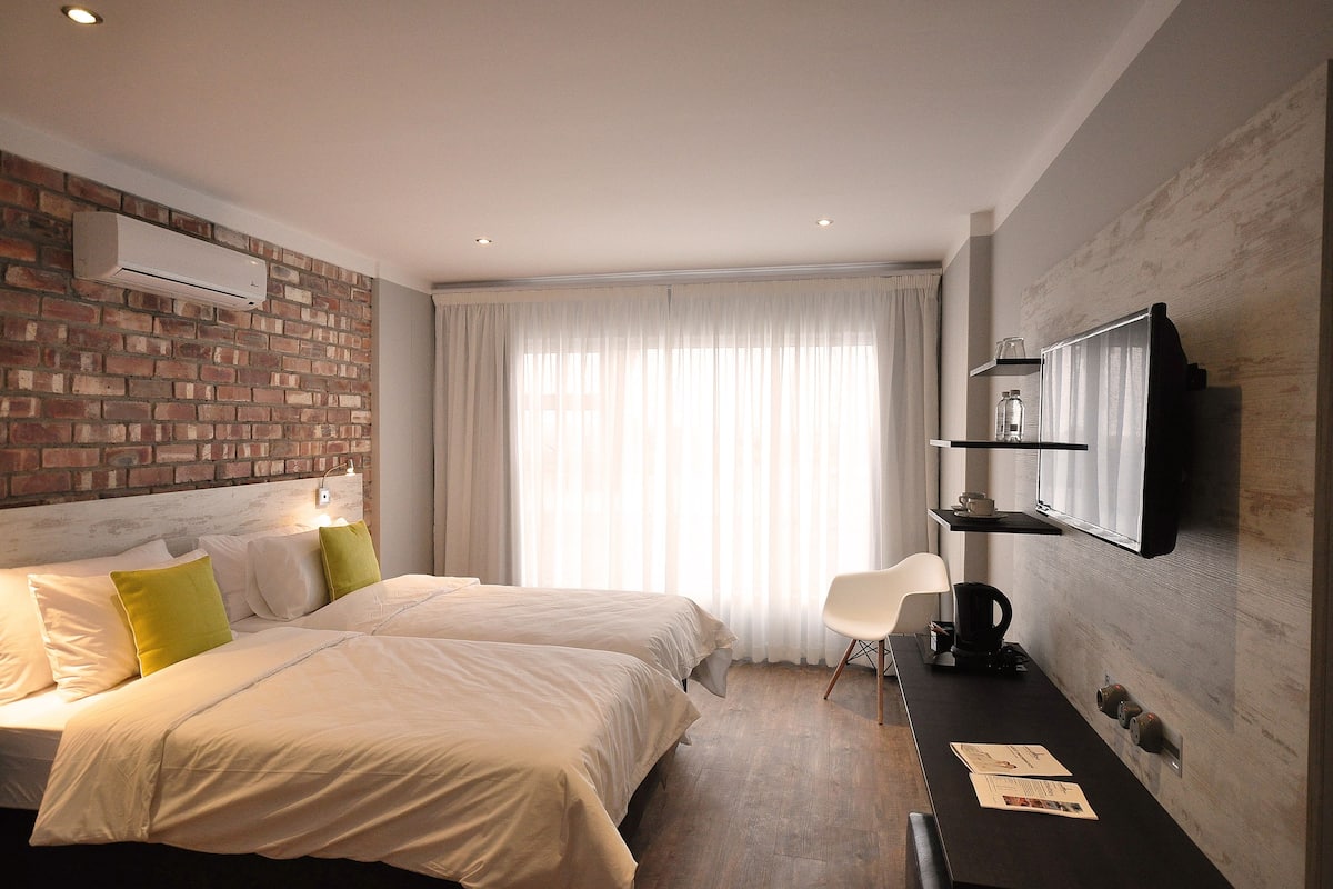 A hotel room with two beds, a flat-screen TV mounted on the wall, a coffee machine, and a brick-patterned wall.