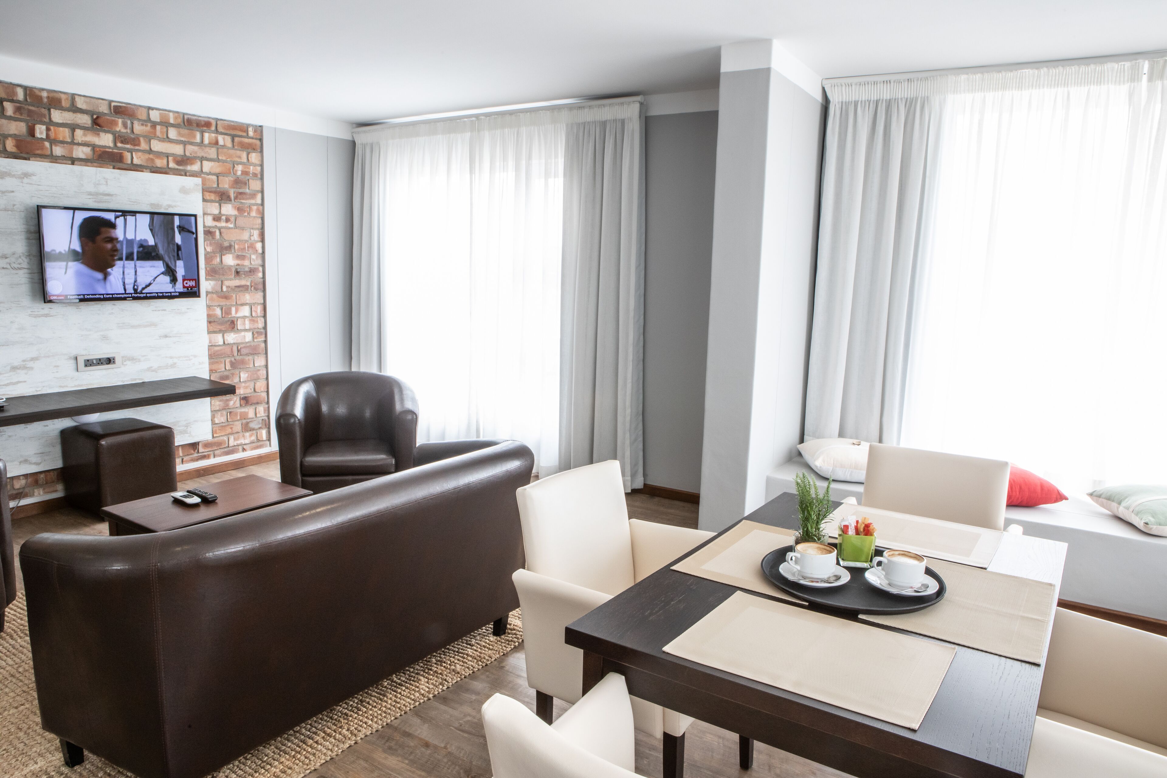 Una habitación de hotel moderna con un sofá de cuero, una mesa de comedor con cafetera y una televisión montada en una pared de ladrillo, además de ventanas grandes con cortinas.