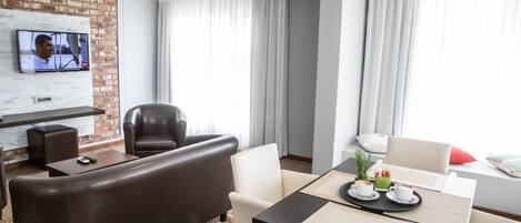 Una habitación de hotel moderna con un sofá de cuero, una mesa de comedor con cafetera y una televisión montada en una pared de ladrillo, además de ventanas grandes con cortinas.
