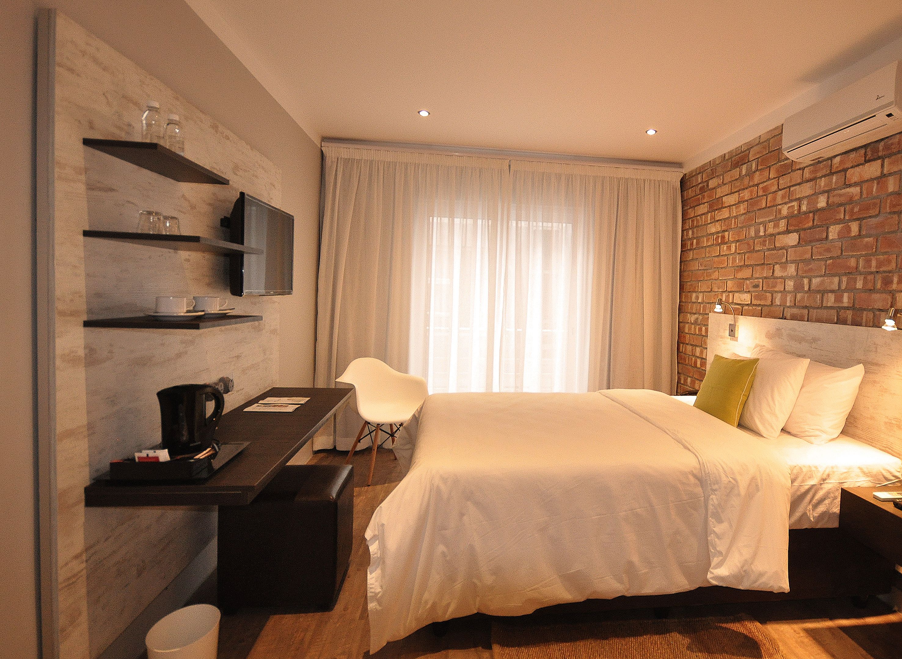 A hotel room with a bed, a desk with a computer, a chair, a TV, and a brick wall.